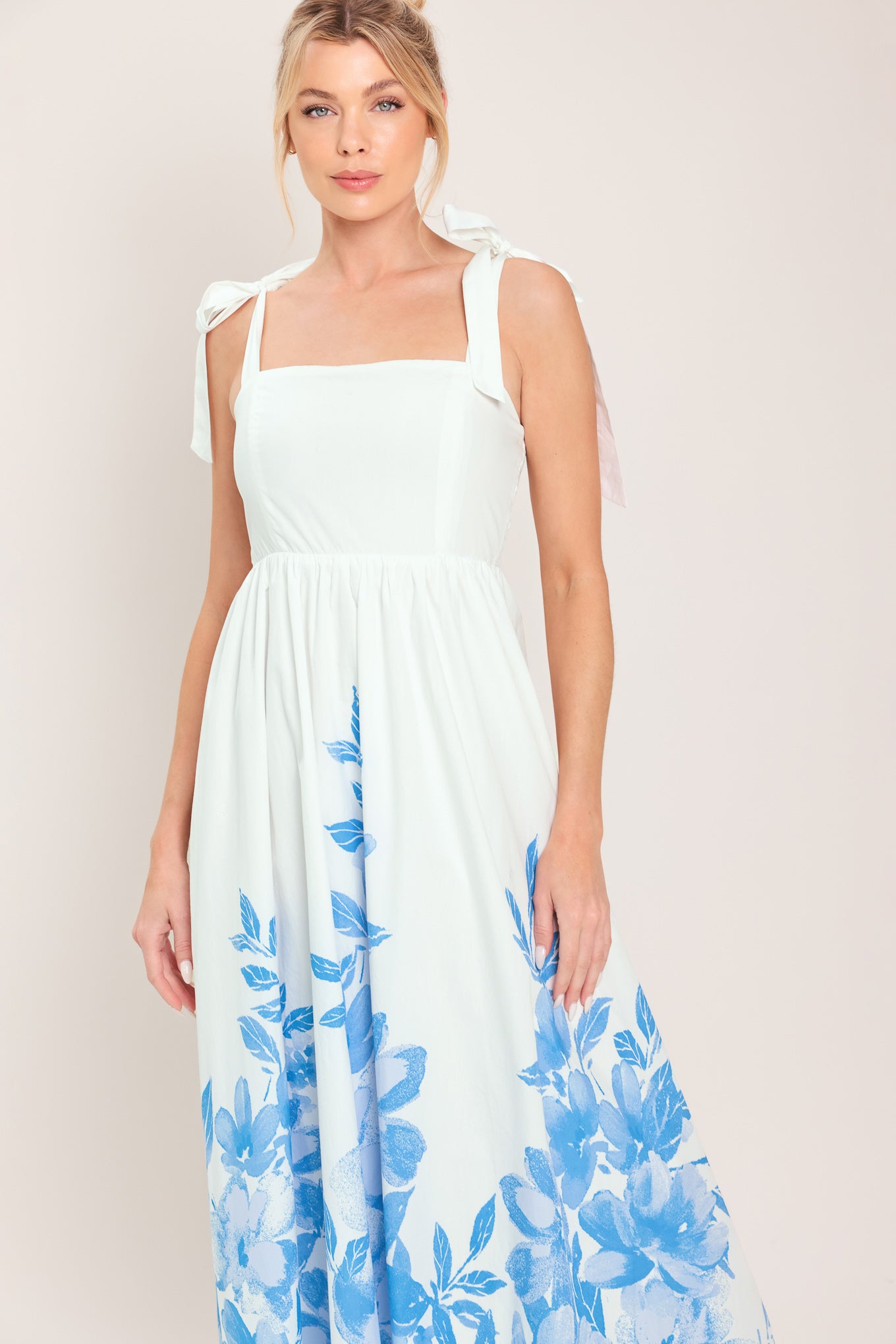 Woman wearing a white maxi dress with blue floral pattern featuring straight neckline, shoulder ties, full skirt and smocked back bodice | Flying Tomato