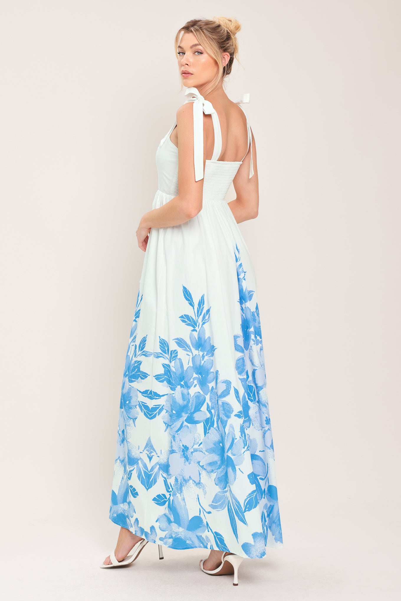 Woman wearing a white maxi dress with blue floral pattern featuring straight neckline, shoulder ties, full skirt and smocked back bodice | Flying Tomato