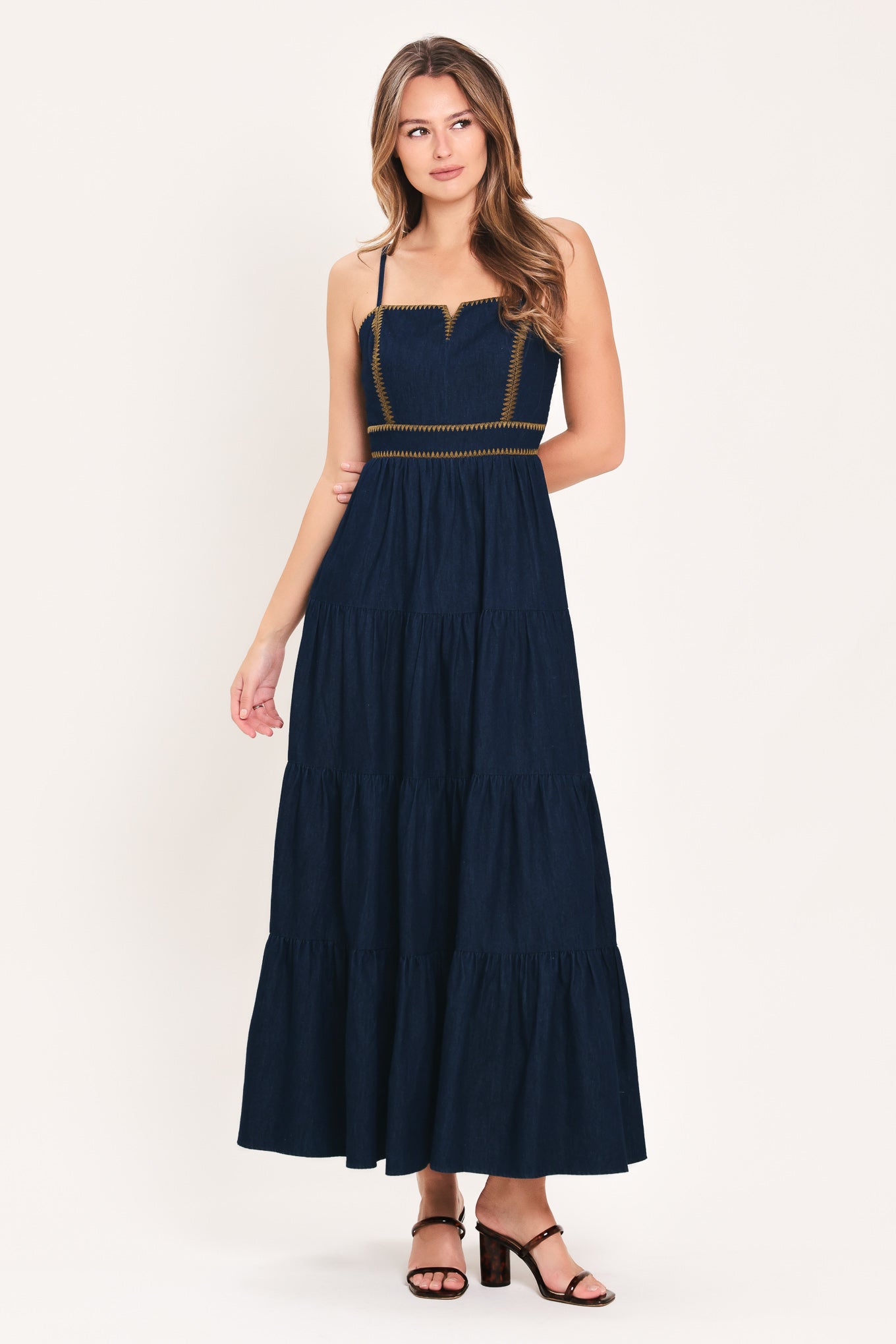 A woman wearing an indigo navy blue denim maxi dress with thin straps, a sweetheart neckline, and gold stitching detailing at the waist | Flying Tomato