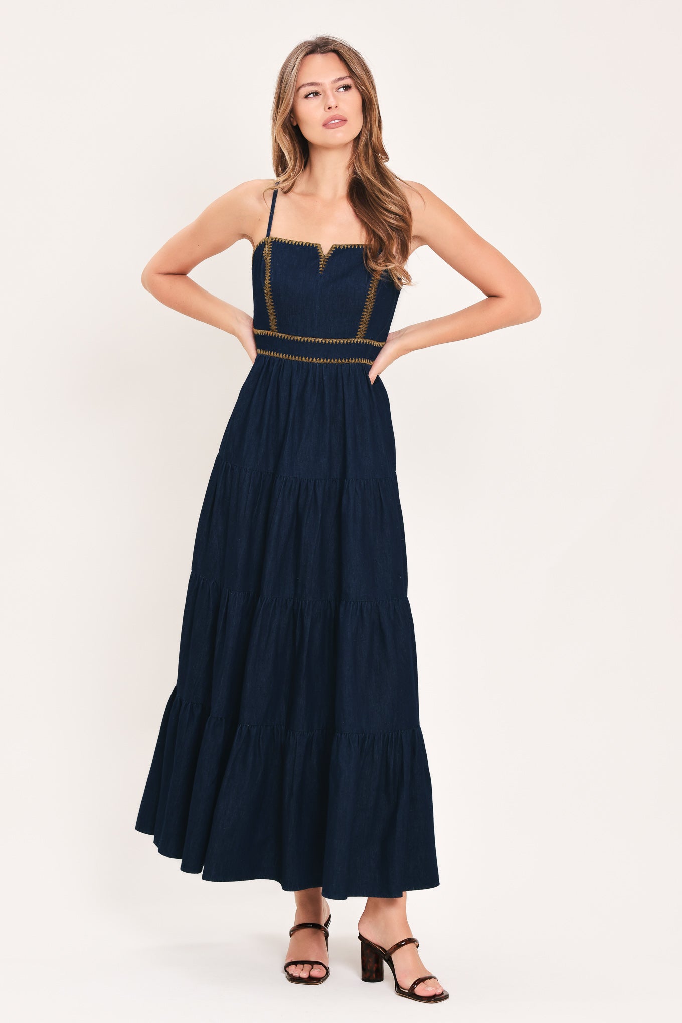 A woman wearing an indigo navy blue denim maxi dress with thin straps, a sweetheart neckline, and gold stitching detailing at the waist | Flying Tomato