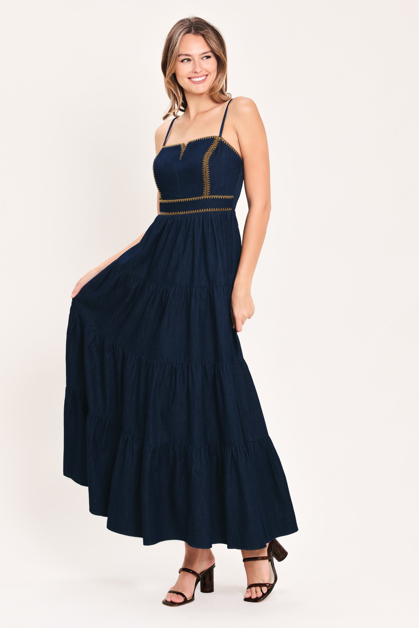 A woman wearing an indigo navy blue denim maxi dress with thin straps, a sweetheart neckline, and gold stitching detailing at the waist | Flying Tomato