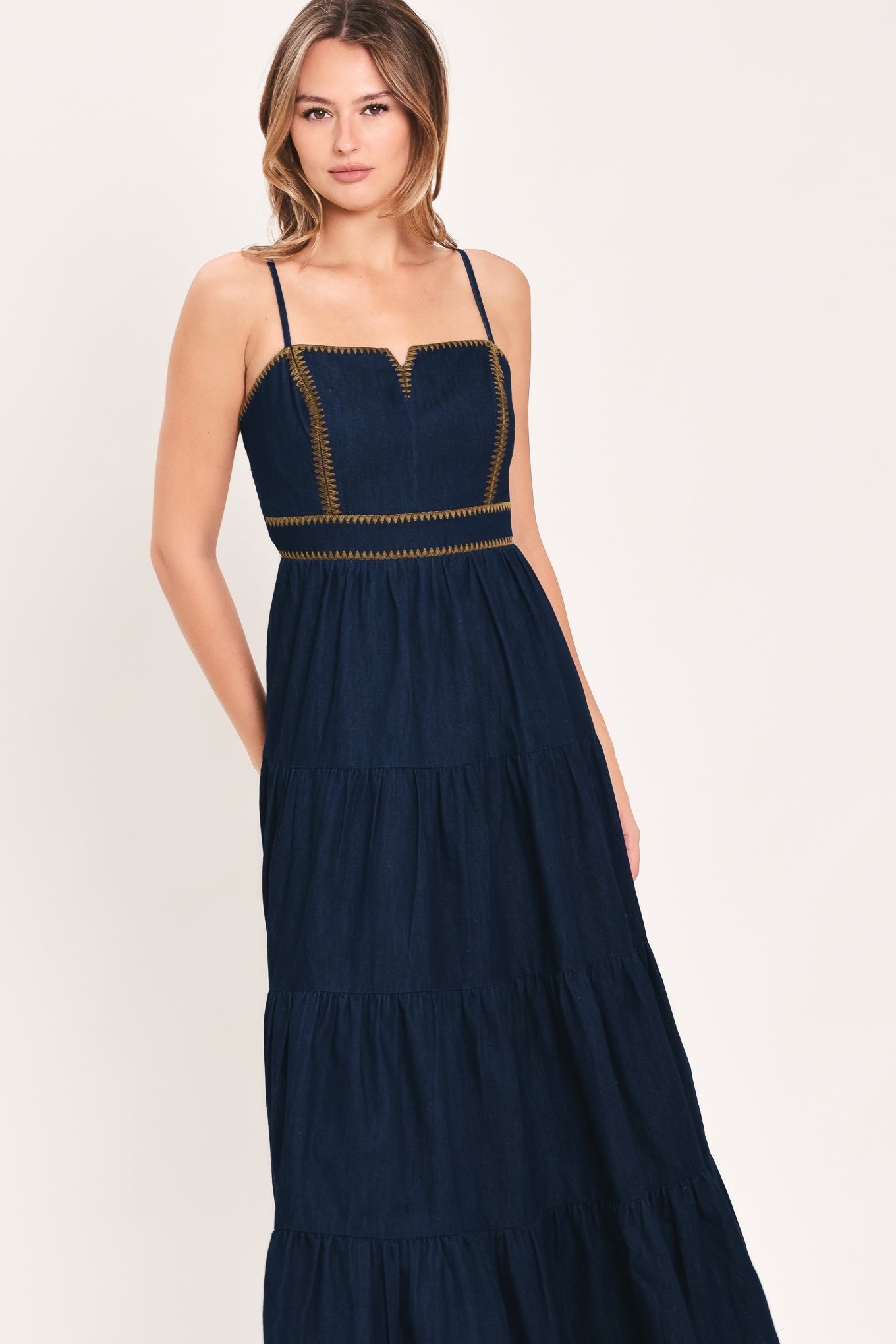 A woman wearing an indigo navy blue denim maxi dress with thin straps, a sweetheart neckline, and gold stitching detailing at the waist | Flying Tomato