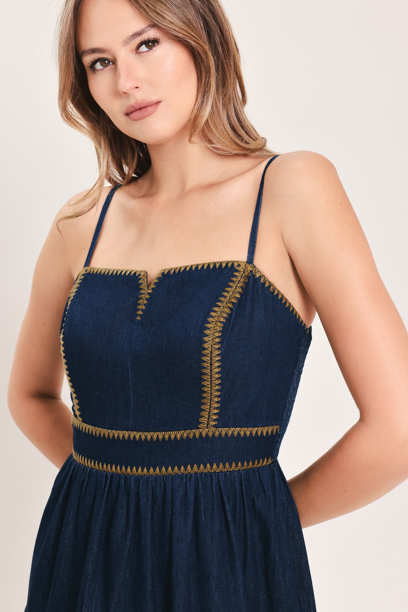 A woman wearing an indigo navy blue denim maxi dress with thin straps, a sweetheart neckline, and gold stitching detailing at the waist | Flying Tomato