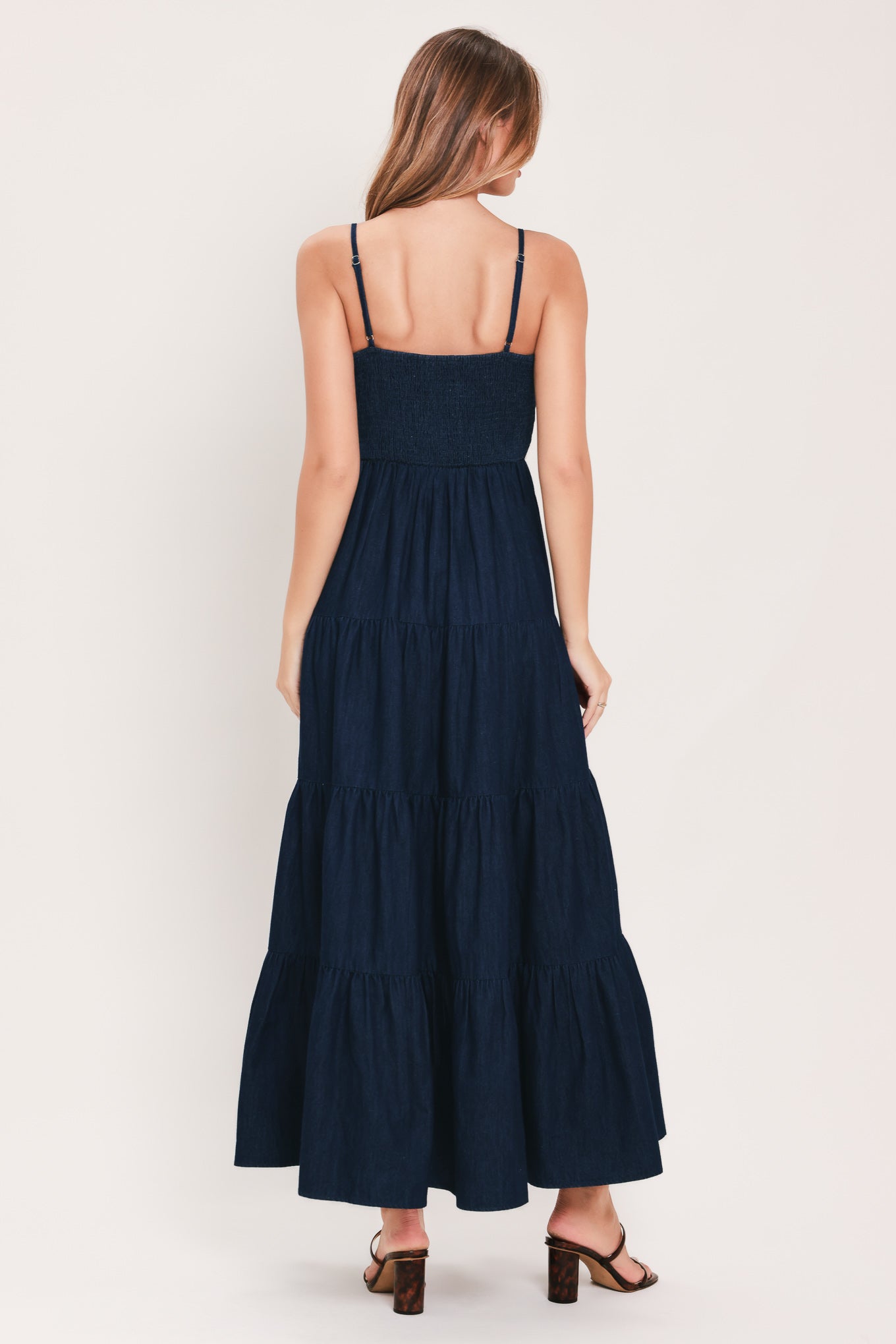 A woman wearing an indigo navy blue denim maxi dress with thin straps, a sweetheart neckline, and gold stitching detailing at the waist | Flying Tomato