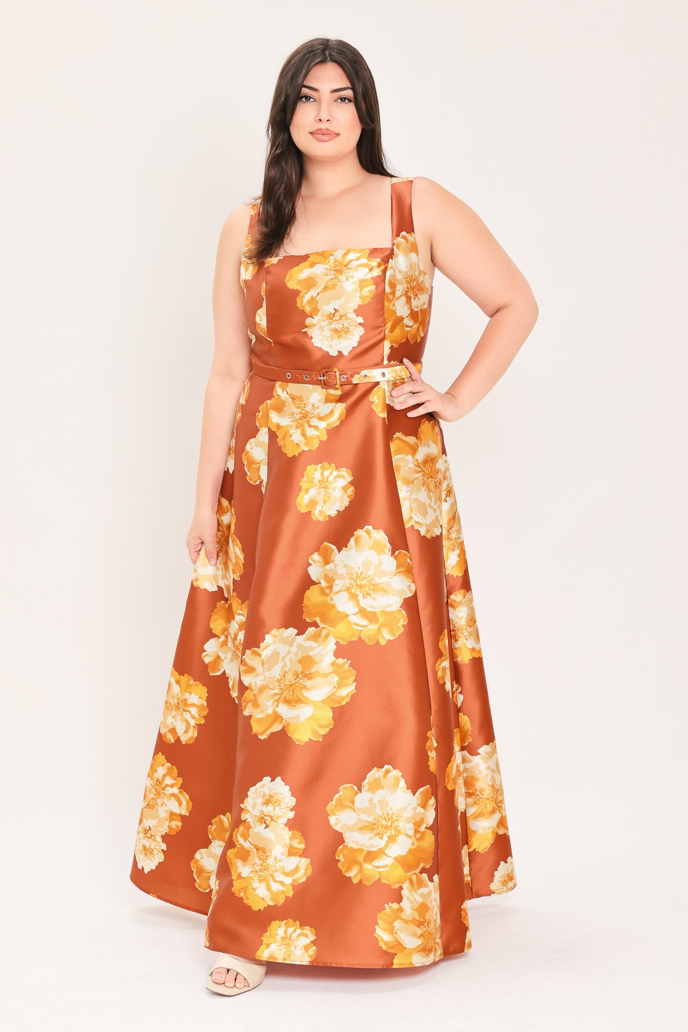 A woman modeling a sleeveless, maxi satin dress in rust orange with a large yellow and white floral print, featuring a matching belt at the waist | Flying Tomato Curve
