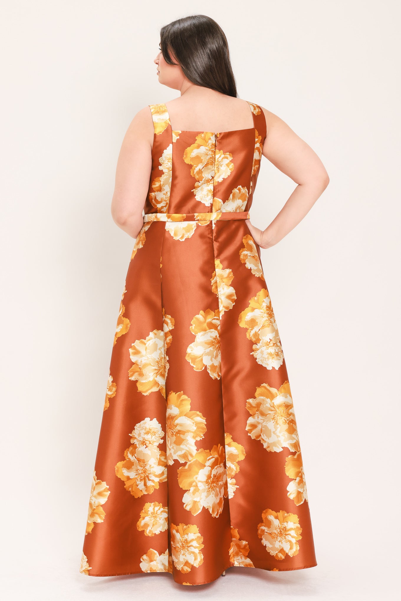 A woman modeling a sleeveless, maxi satin dress in rust orange with a large yellow and white floral print, featuring a matching belt at the waist | Flying Tomato Curve