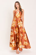 A woman modeling a sleeveless, maxi satin dress in rust orange with a large yellow and white floral print, featuring a matching belt at the waist | Flying Tomato