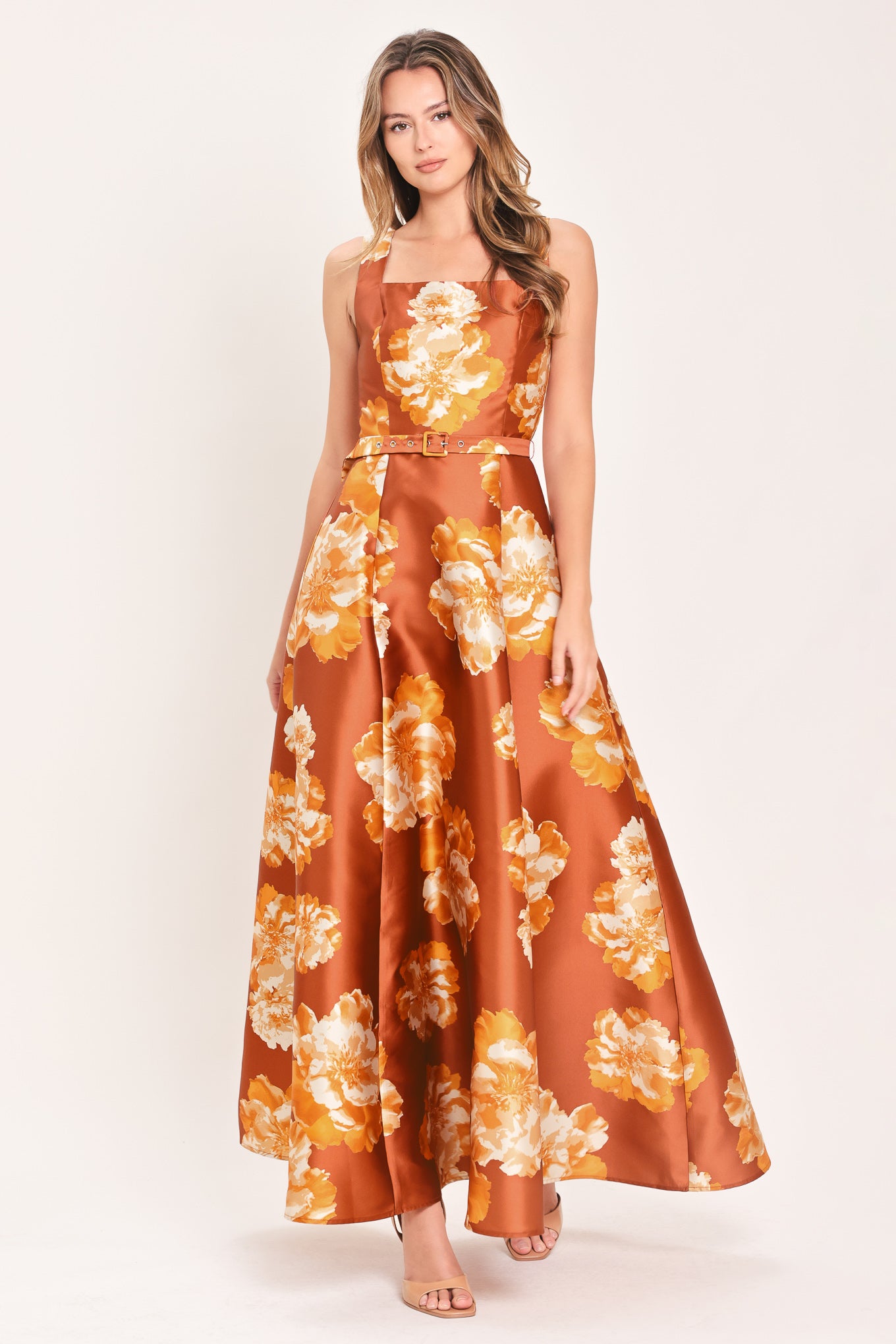 A woman modeling a sleeveless, maxi satin dress in rust orange with a large yellow and white floral print, featuring a matching belt at the waist | Flying Tomato