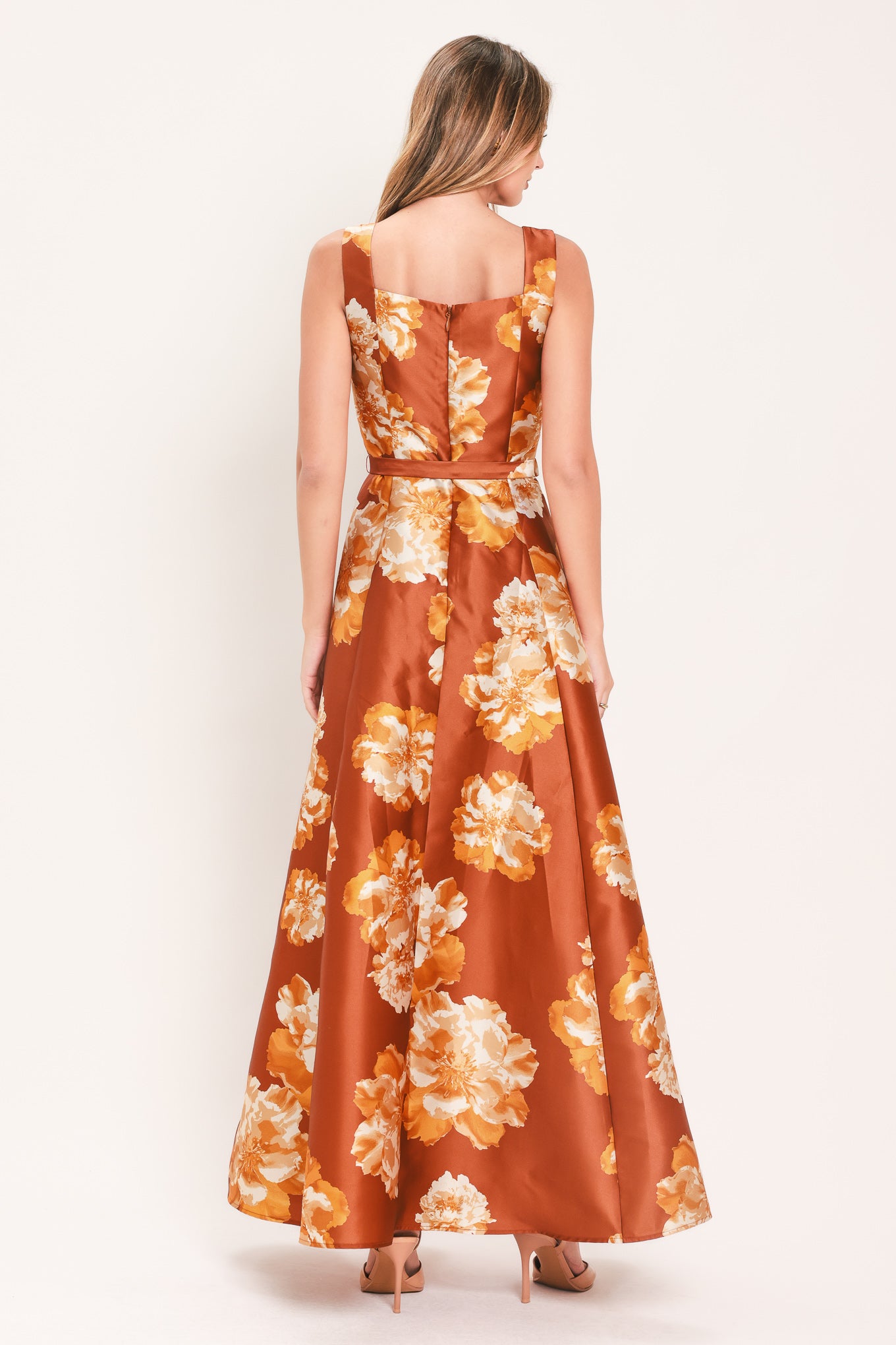 A woman modeling a sleeveless, maxi satin dress in rust orange with a large yellow and white floral print, featuring a matching belt at the waist | Flying Tomato