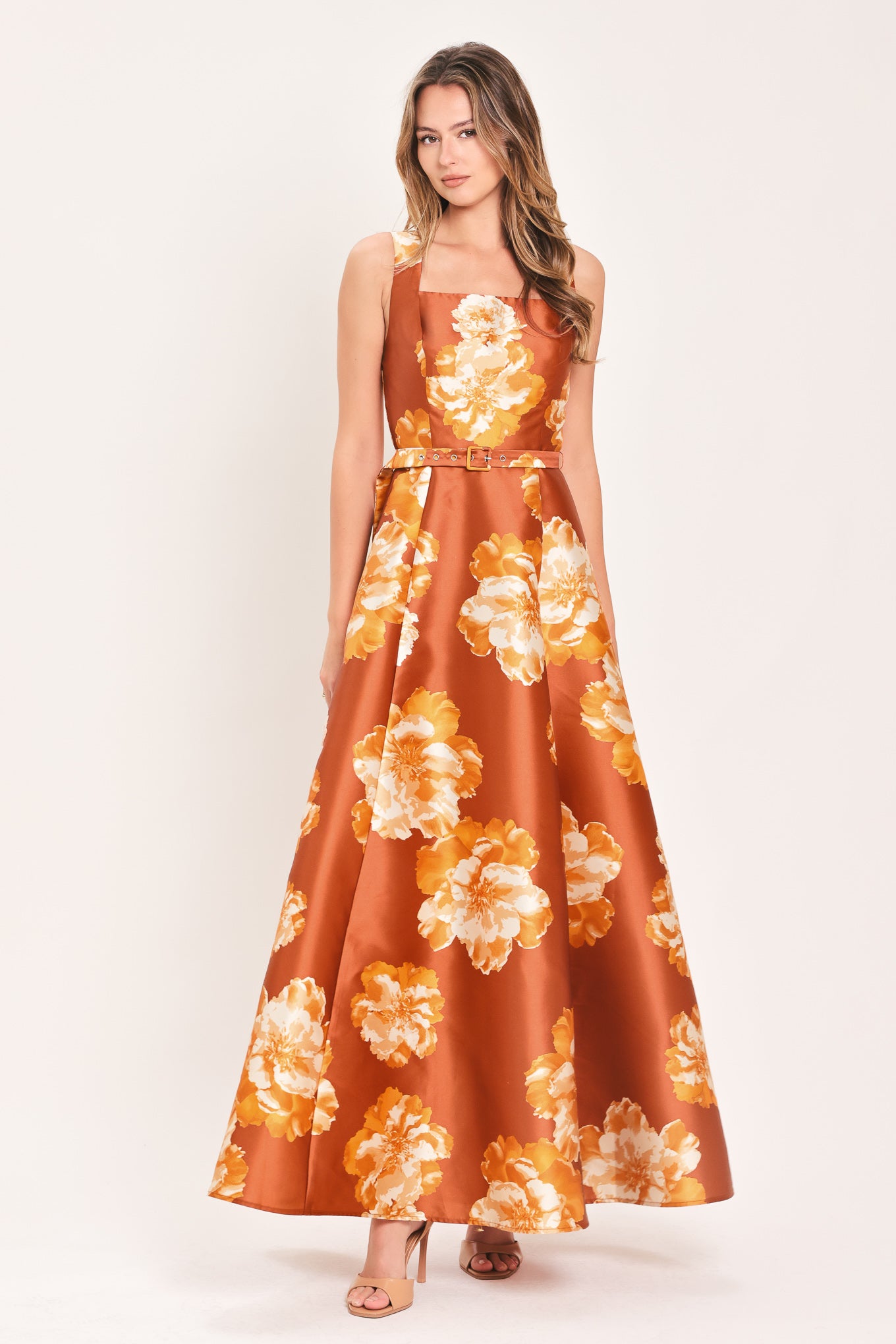 A woman modeling a sleeveless, maxi satin dress in rust orange with a large yellow and white floral print, featuring a matching belt at the waist | Flying Tomato