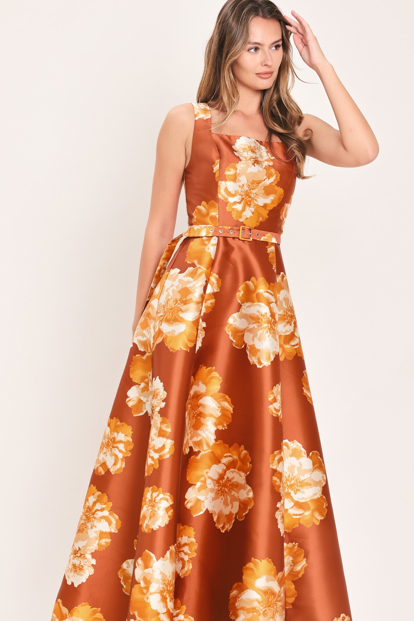 A woman modeling a sleeveless, maxi satin dress in rust orange with a large yellow and white floral print, featuring a matching belt at the waist | Flying Tomato