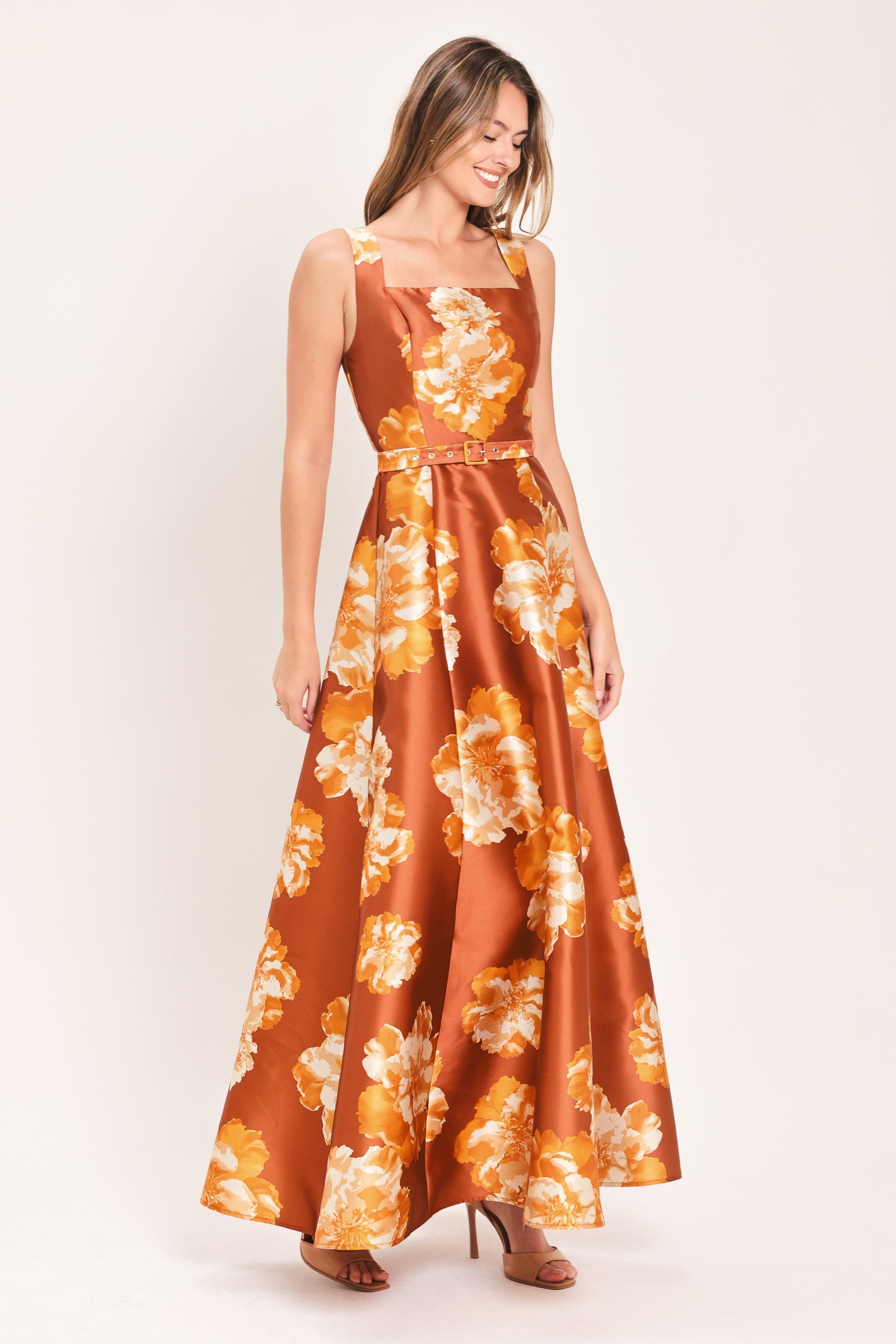 A woman modeling a sleeveless, maxi satin dress in rust orange with a large yellow and white floral print, featuring a matching belt at the waist | Flying Tomato
