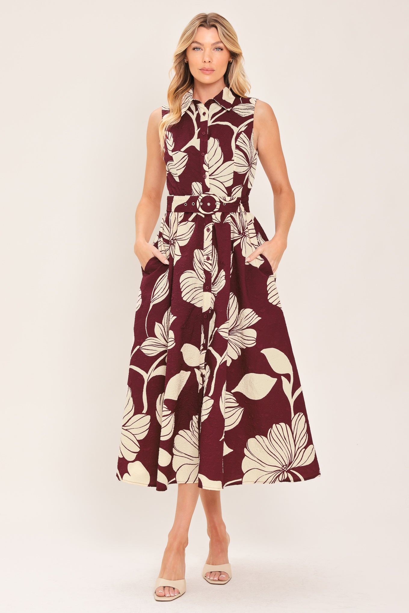 A woman wearing a sleeveless burgundy midi dress with a deep V-neck, a matching fabric tie belt with a decorative center medallion, and a bold cream floral print | Flying Tomato