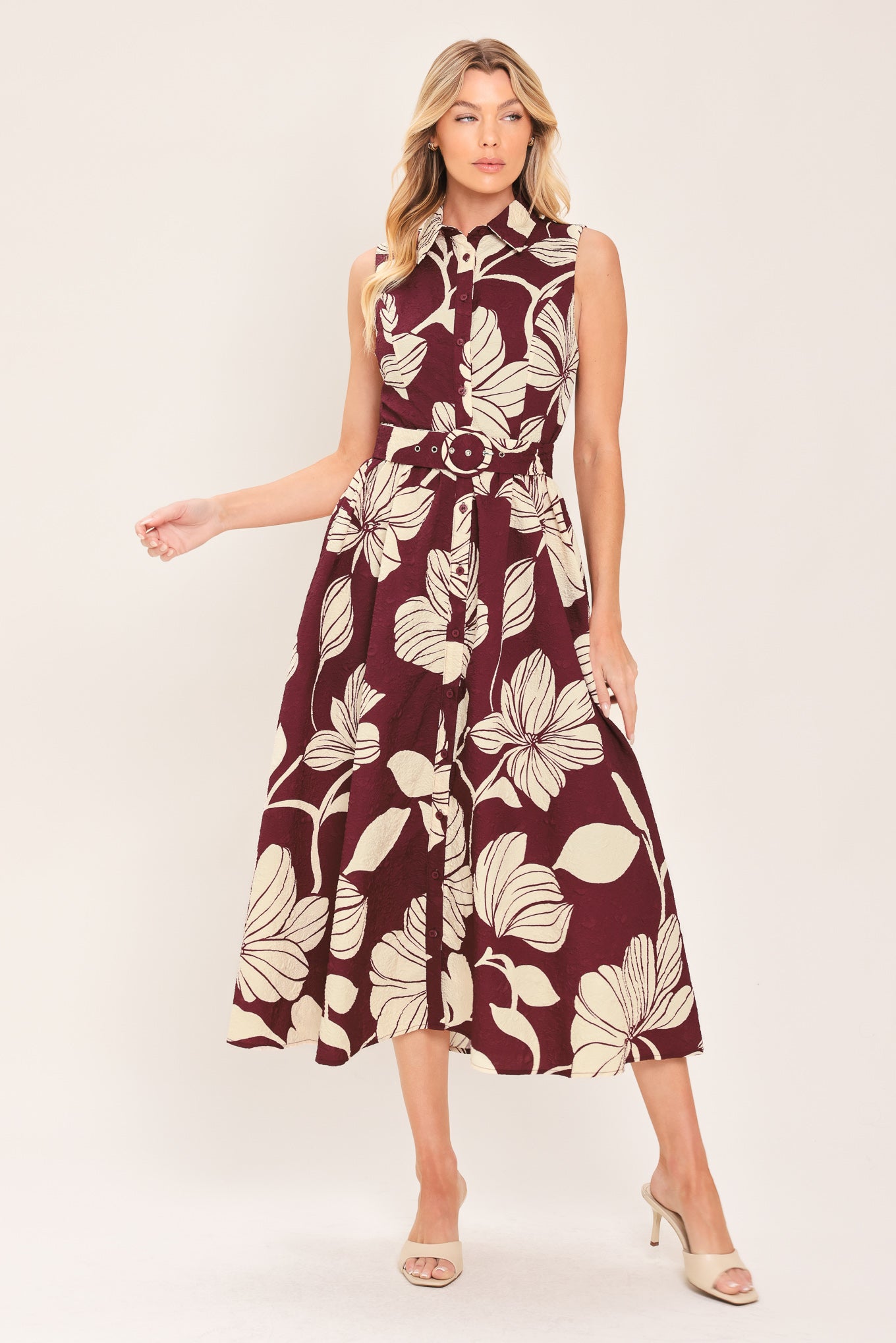 A woman wearing a sleeveless burgundy midi dress with a deep V-neck, a matching fabric tie belt with a decorative center medallion, and a bold cream floral print | Flying Tomato