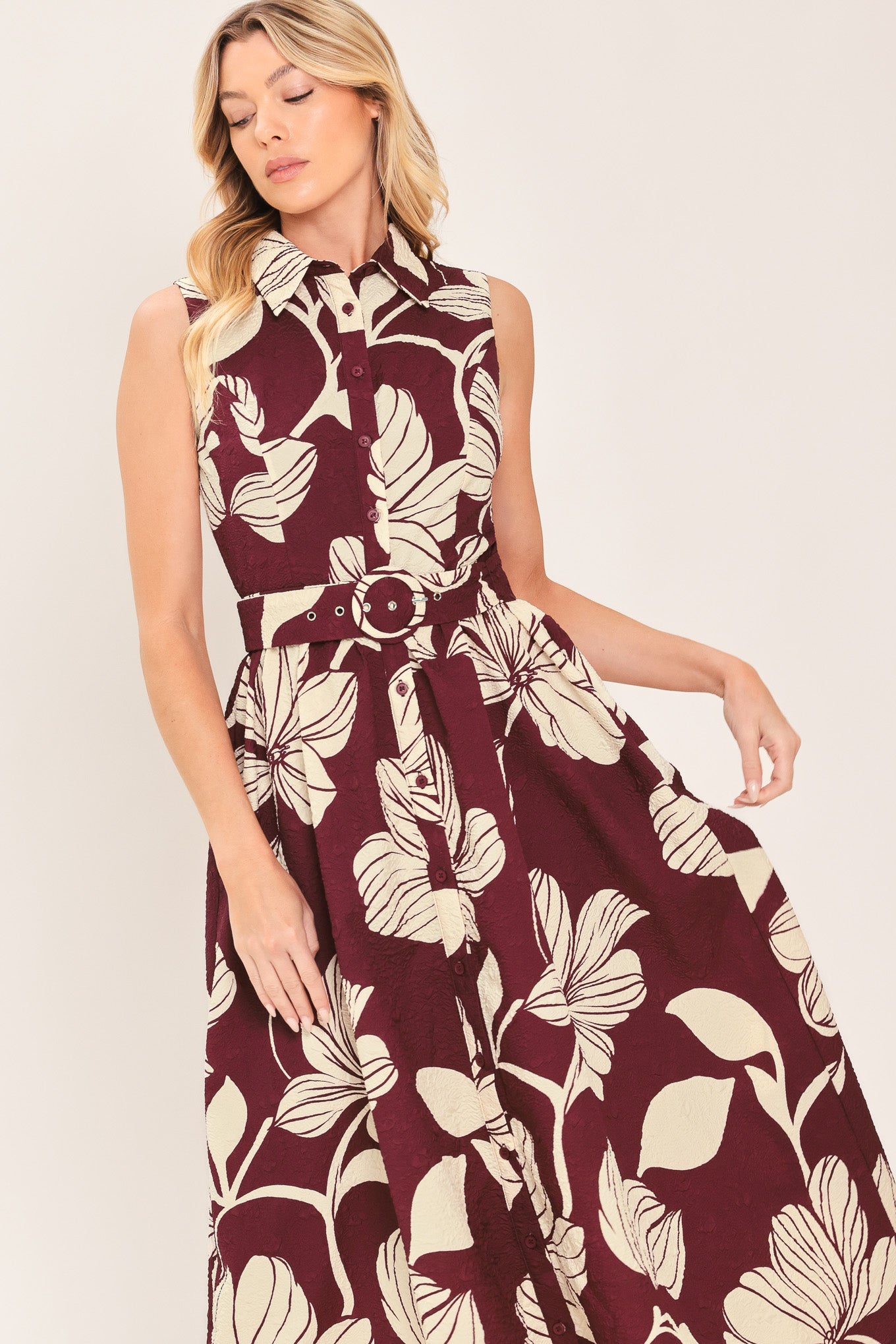 A woman wearing a sleeveless burgundy midi dress with a deep V-neck, a matching fabric tie belt with a decorative center medallion, and a bold cream floral print | Flying Tomato