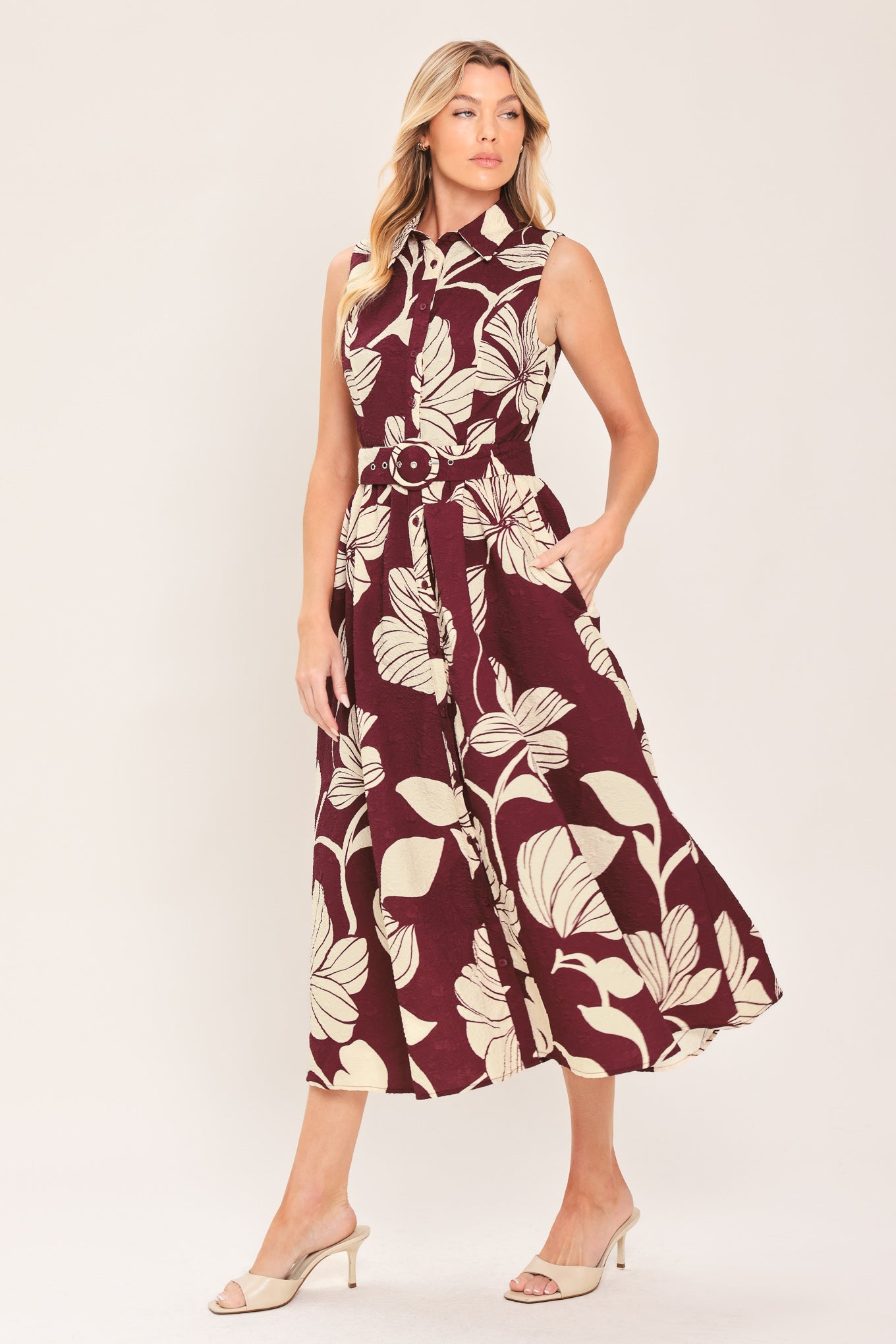A woman wearing a sleeveless burgundy midi dress with a deep V-neck, a matching fabric tie belt with a decorative center medallion, and a bold cream floral print | Flying Tomato