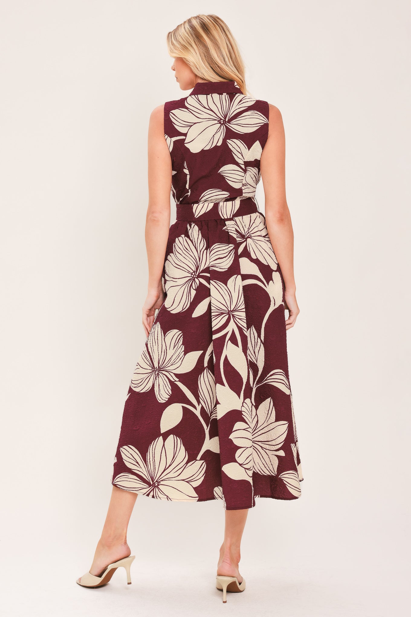 A woman wearing a sleeveless burgundy midi dress with a deep V-neck, a matching fabric tie belt with a decorative center medallion, and a bold cream floral print | Flying Tomato