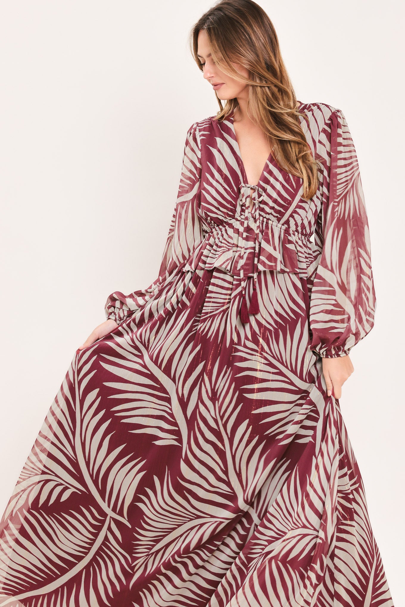 A woman wearing a long-sleeve maxi dress with a deep V-neck and a tropical palm leaf print in burgundy and cream | Flying Tomato