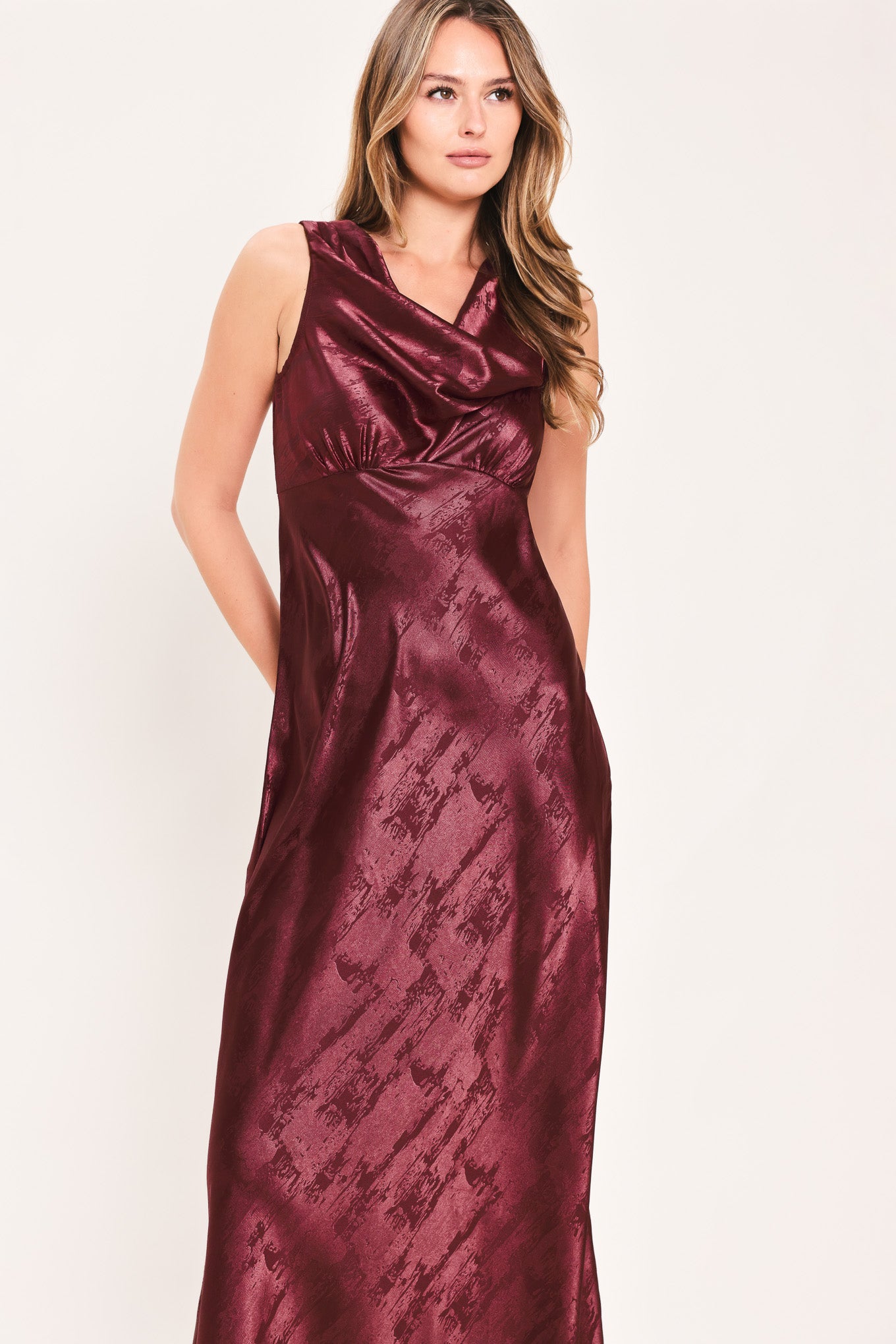 A woman wearing a sleeveless, floor-length metallic burgundy gown with a draped neckline and a subtle crinkle texture | Flying Tomato