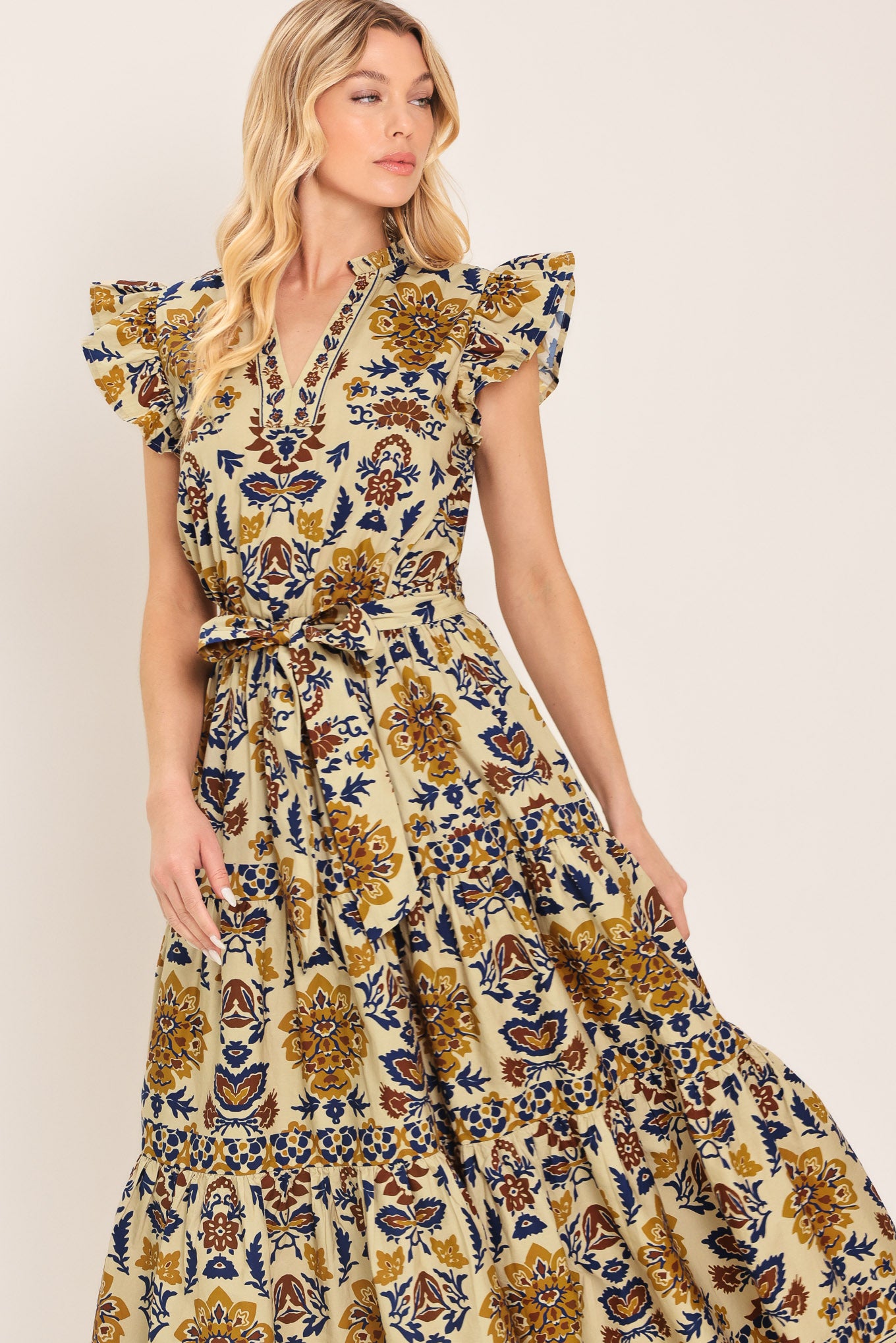 Woman wearing a taupe printed midi dress featuring V neckline with ruffled edge, short ruffle sleeve, elastic waist, self sash and tier skirt | Flying Tomato