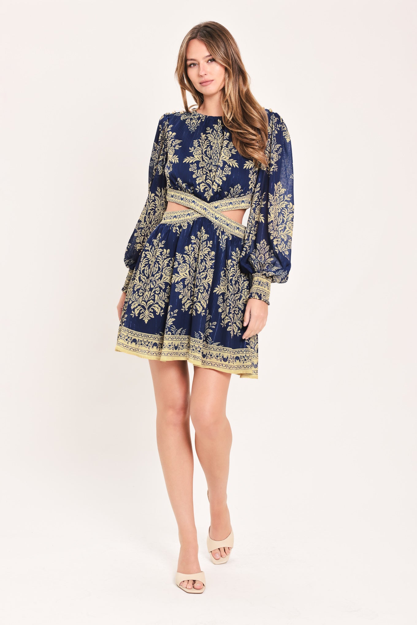 A woman wearing a navy blue long-sleeve mini dress with a beige and gold botanical pattern, a waist cutout, and a flared skirt | Flying Tomato