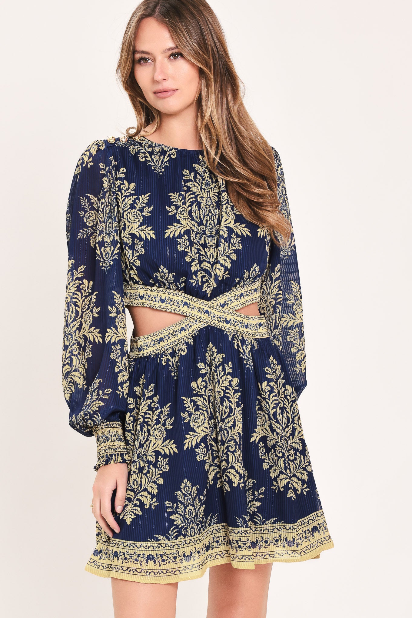 A woman wearing a navy blue long-sleeve mini dress with a beige and gold botanical pattern, a waist cutout, and a flared skirt | Flying Tomato