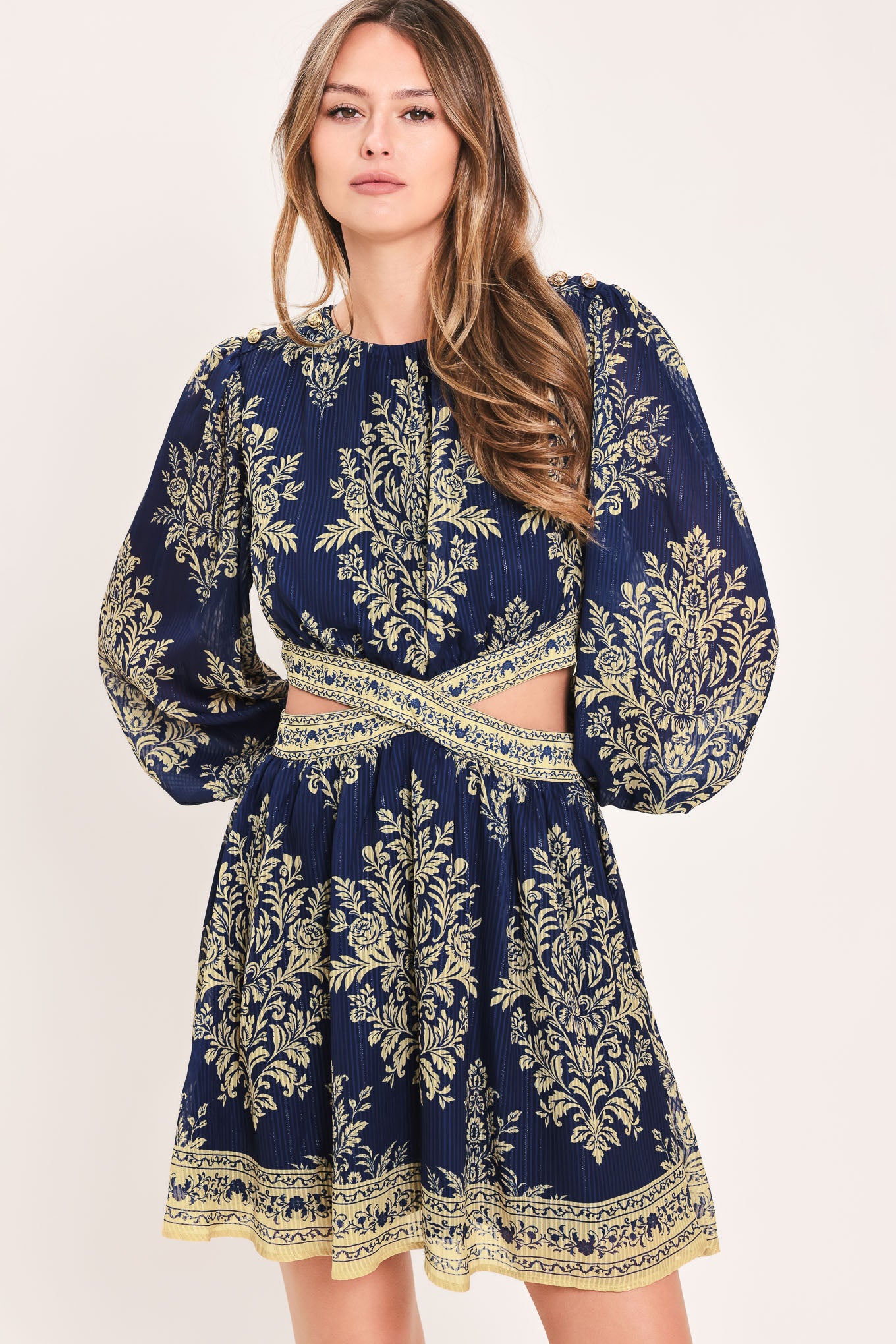 A woman wearing a navy blue long-sleeve mini dress with a beige and gold botanical pattern, a waist cutout, and a flared skirt | Flying Tomato