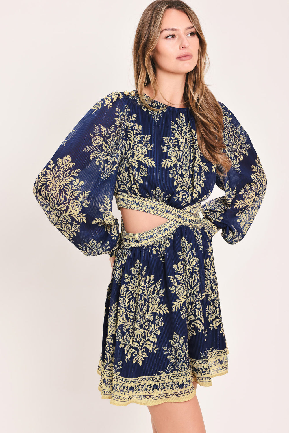 A woman wearing a navy blue long-sleeve mini dress with a beige and gold botanical pattern, a waist cutout, and a flared skirt | Flying Tomato