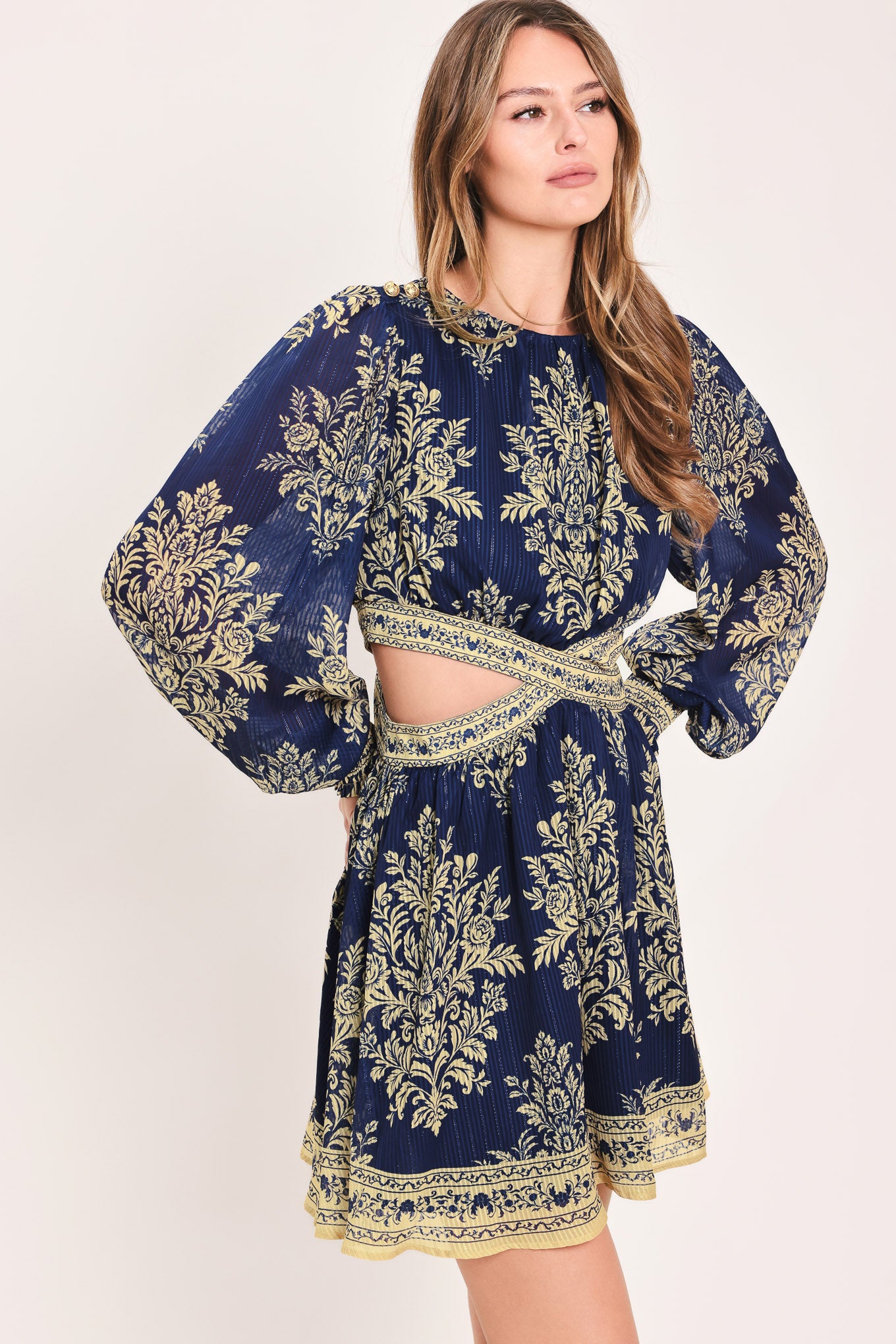 A woman wearing a navy blue long-sleeve mini dress with a beige and gold botanical pattern, a waist cutout, and a flared skirt | Flying Tomato