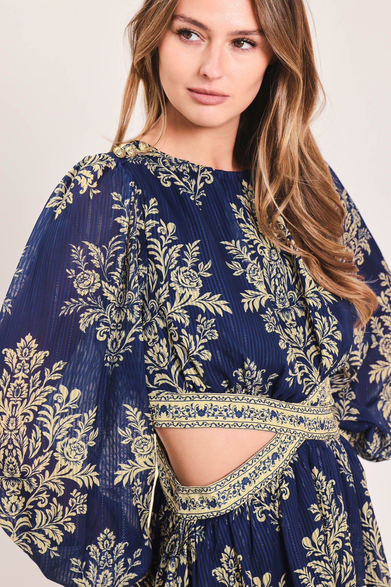 A woman wearing a navy blue long-sleeve mini dress with a beige and gold botanical pattern, a waist cutout, and a flared skirt | Flying Tomato