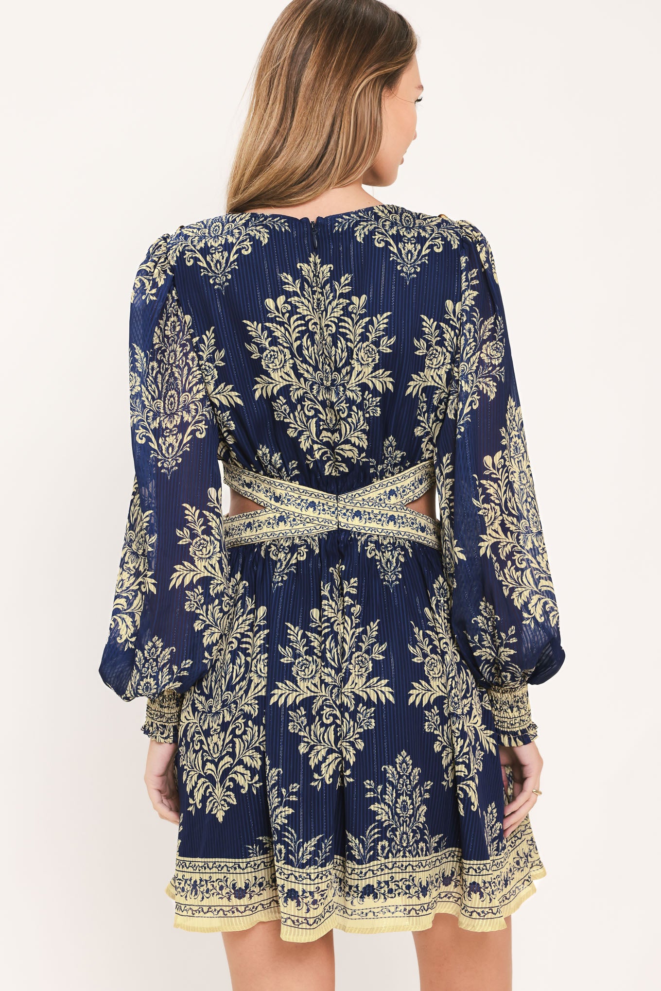 A woman wearing a navy blue long-sleeve mini dress with a beige and gold botanical pattern, a waist cutout, and a flared skirt | Flying Tomato
