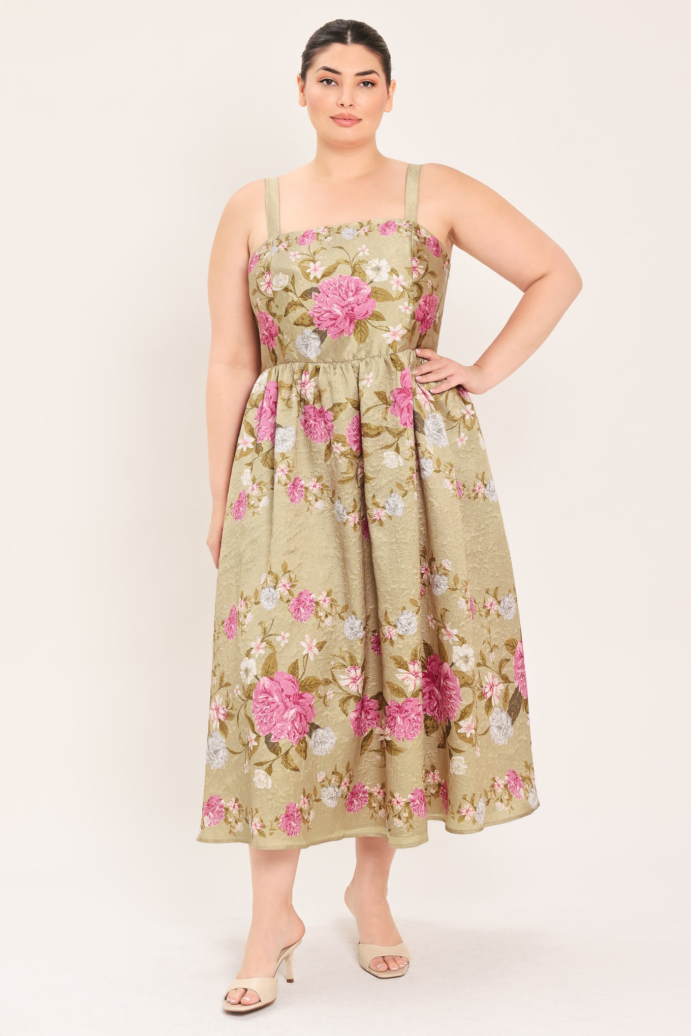 A woman modeling a sleeveless midi dress with a floral print on a muted tan background. The dress features pink, white, and green flowers with a gathered waist | Curve Plus Flying Tomato