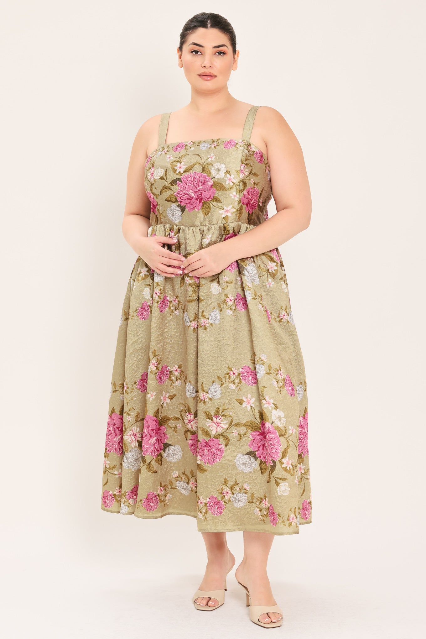 A woman modeling a sleeveless midi dress with a floral print on a muted tan background. The dress features pink, white, and green flowers with a gathered waist | Curve Plus Flying Tomato