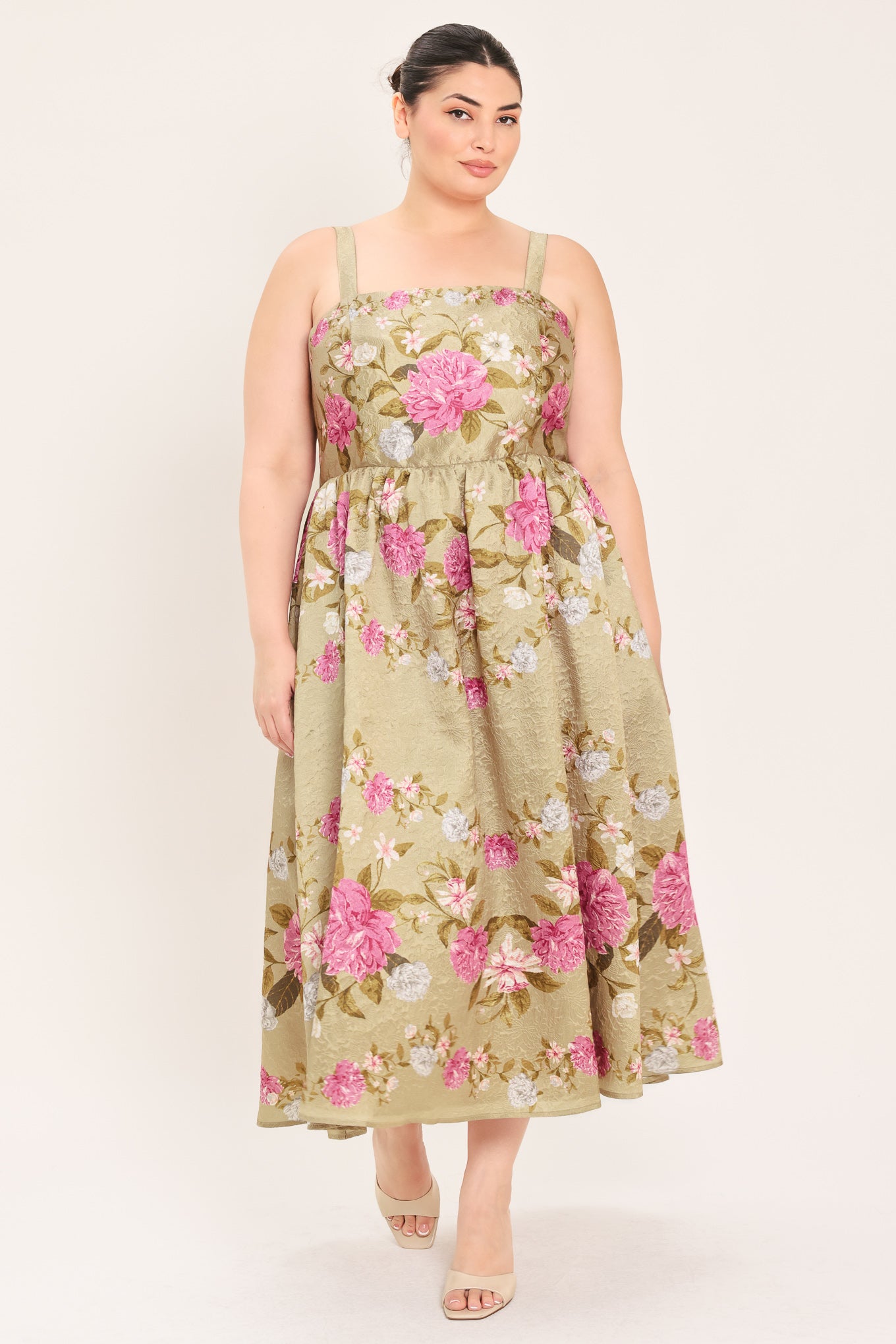 A woman modeling a sleeveless midi dress with a floral print on a muted tan background. The dress features pink, white, and green flowers with a gathered waist | Curve Plus Flying Tomato