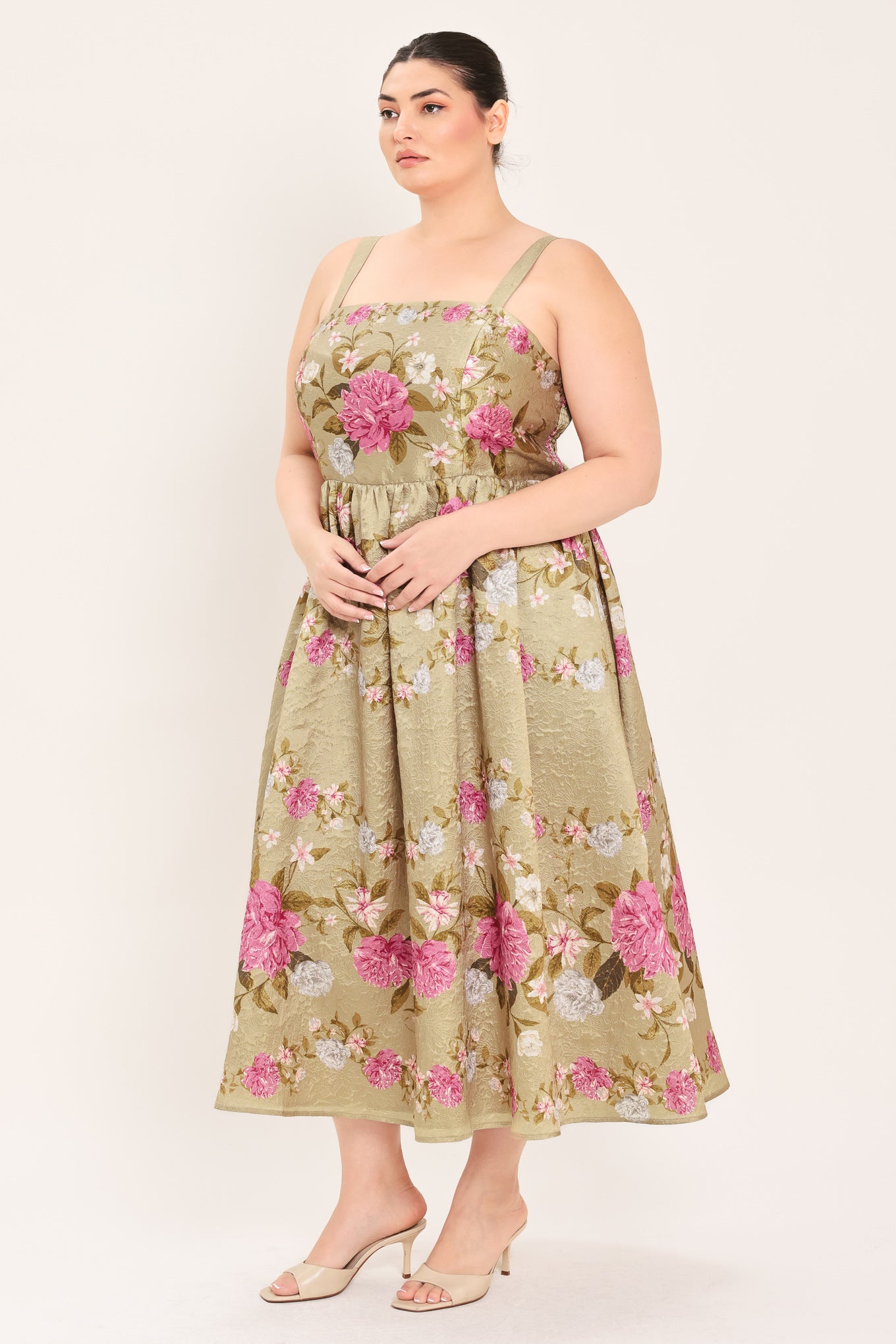 A woman modeling a sleeveless midi dress with a floral print on a muted tan background. The dress features pink, white, and green flowers with a gathered waist | Curve Plus Flying Tomato