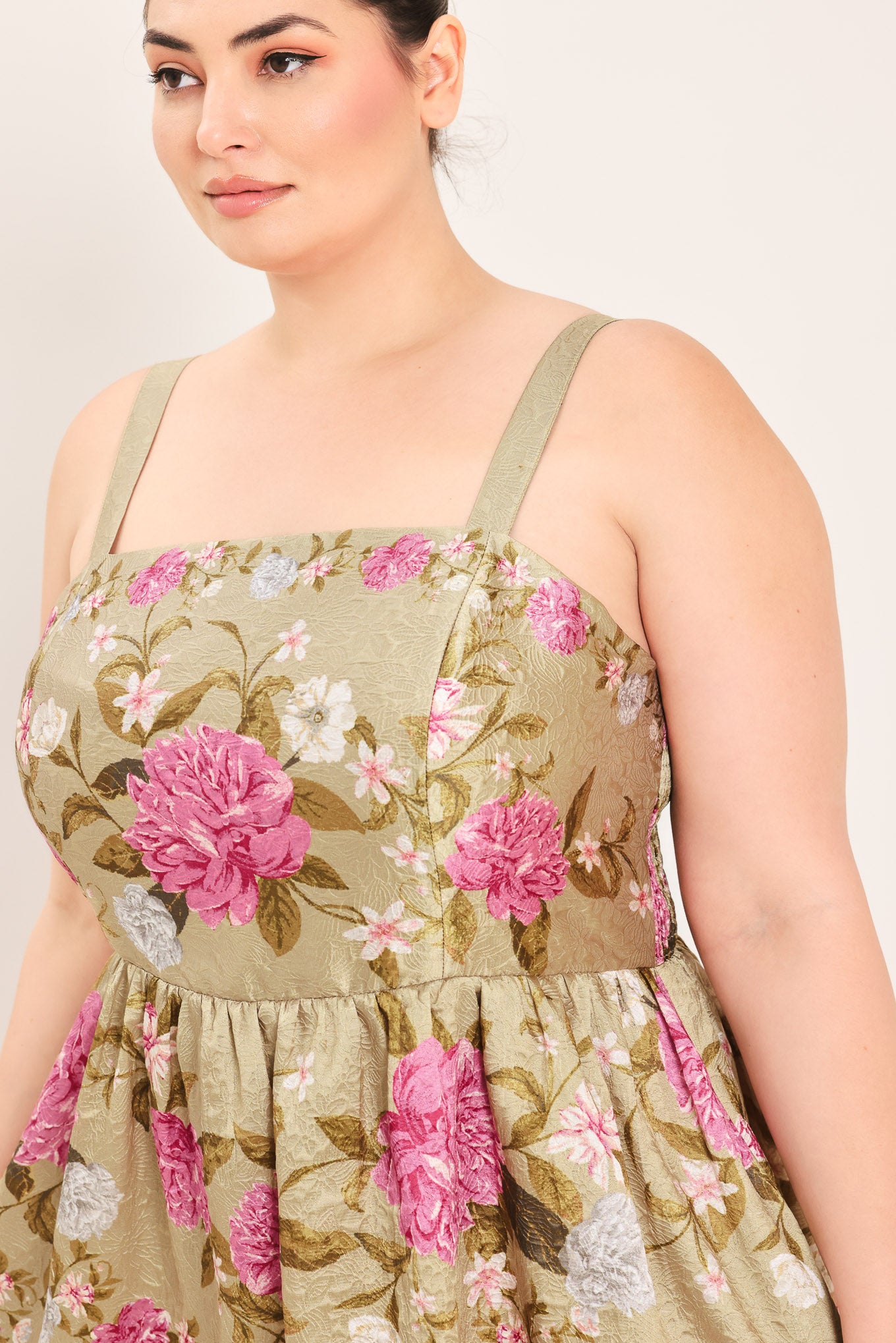 A woman modeling a sleeveless midi dress with a floral print on a muted tan background. The dress features pink, white, and green flowers with a gathered waist | Curve Plus Flying Tomato