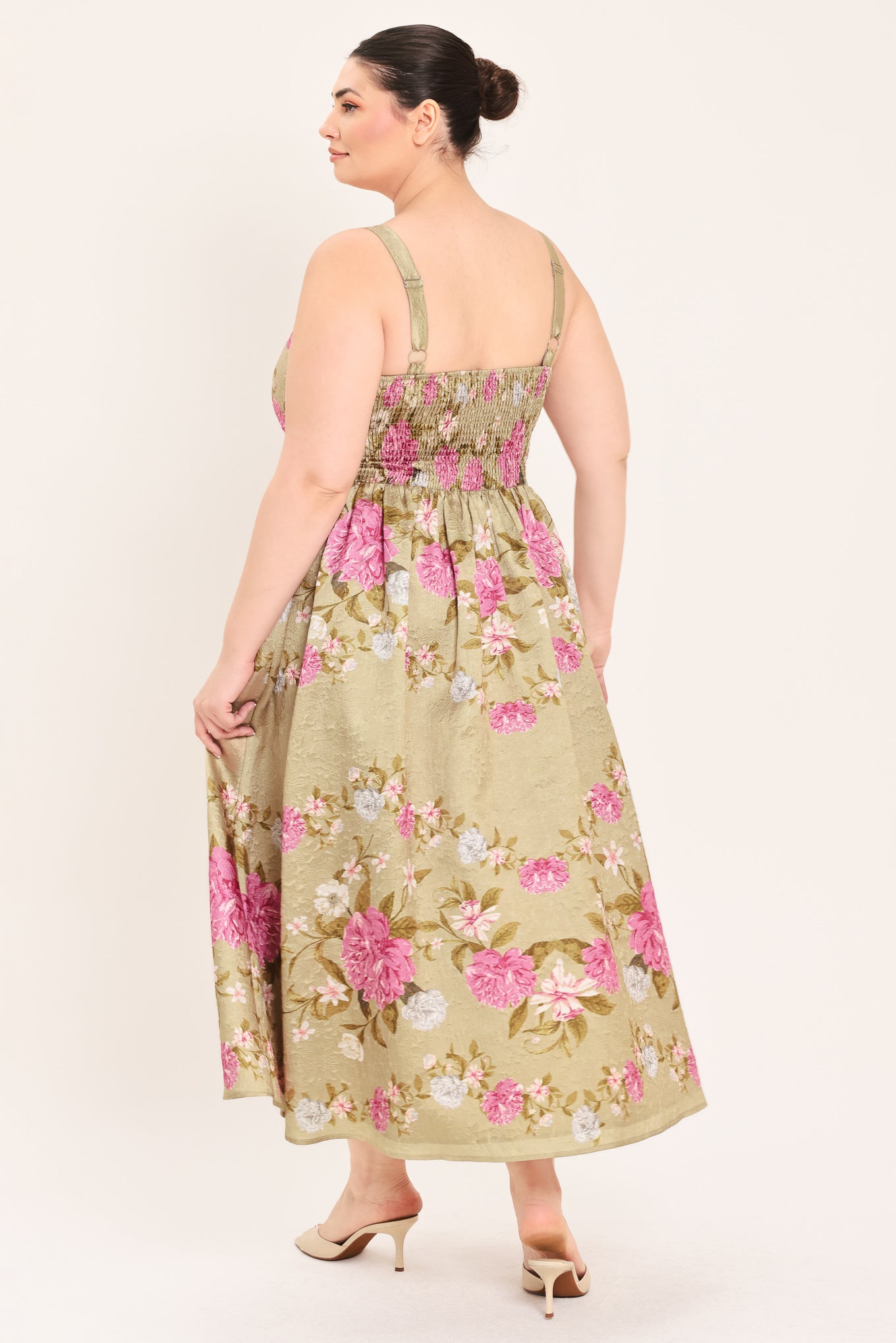 A woman modeling a sleeveless midi dress with a floral print on a muted tan background. The dress features pink, white, and green flowers with a gathered waist | Curve Plus Flying Tomato