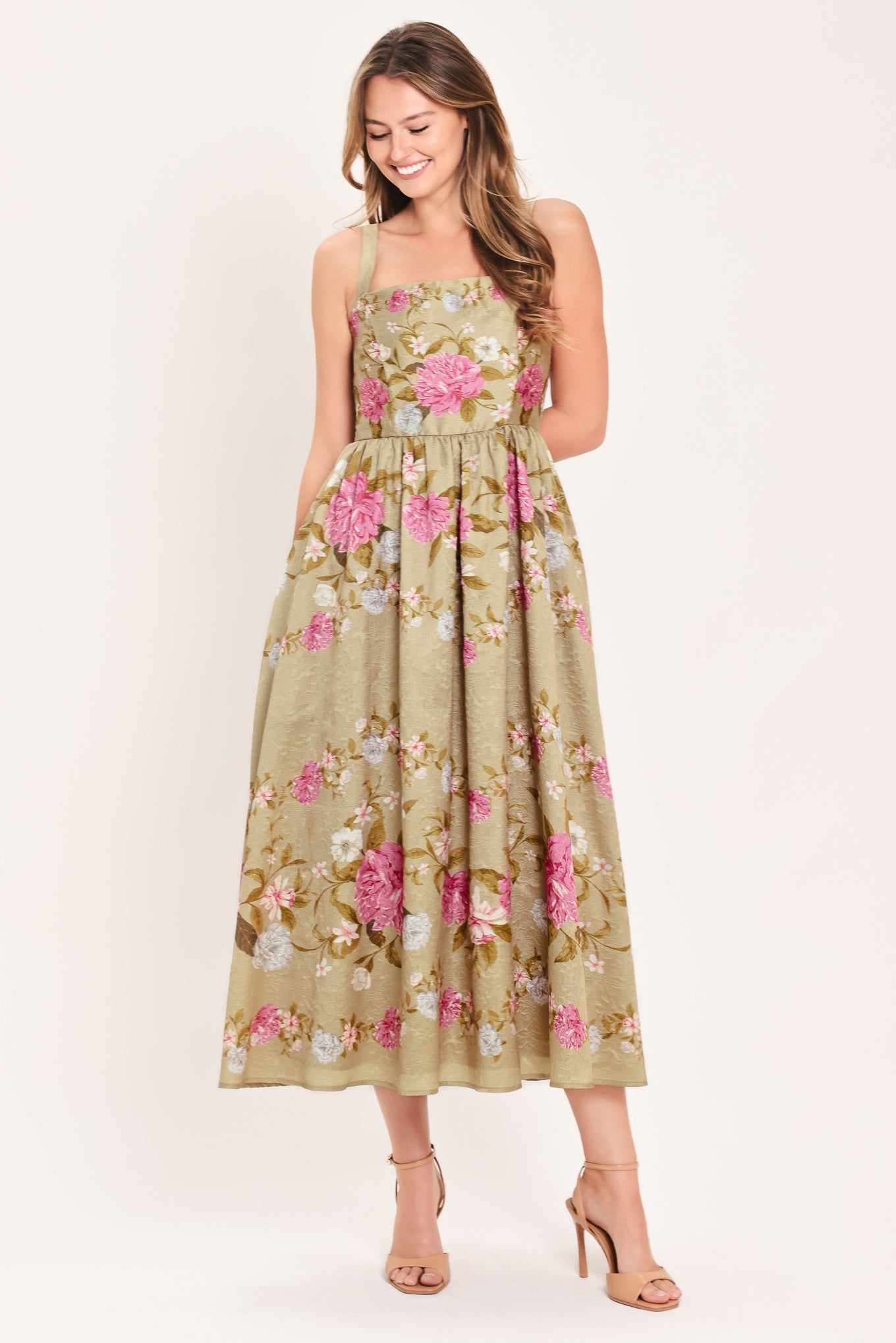 A woman wearing a sleeveless midi dress with a floral print on tan background. The dress features pink, white, and yellow flowers with green leaves | Flying Tomato