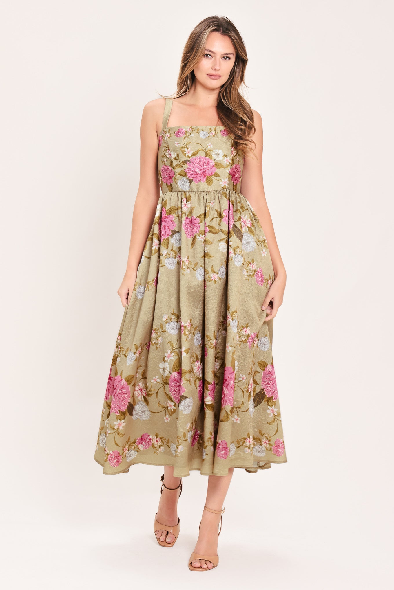A woman wearing a sleeveless midi dress with a floral print on tan background. The dress features pink, white, and yellow flowers with green leaves | Flying Tomato