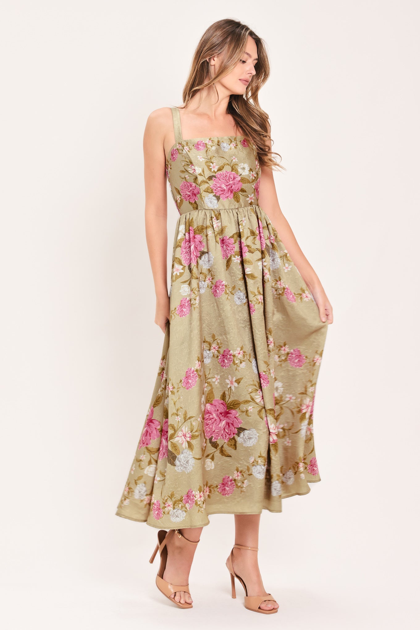 A woman wearing a sleeveless midi dress with a floral print on tan background. The dress features pink, white, and yellow flowers with green leaves | Flying Tomato