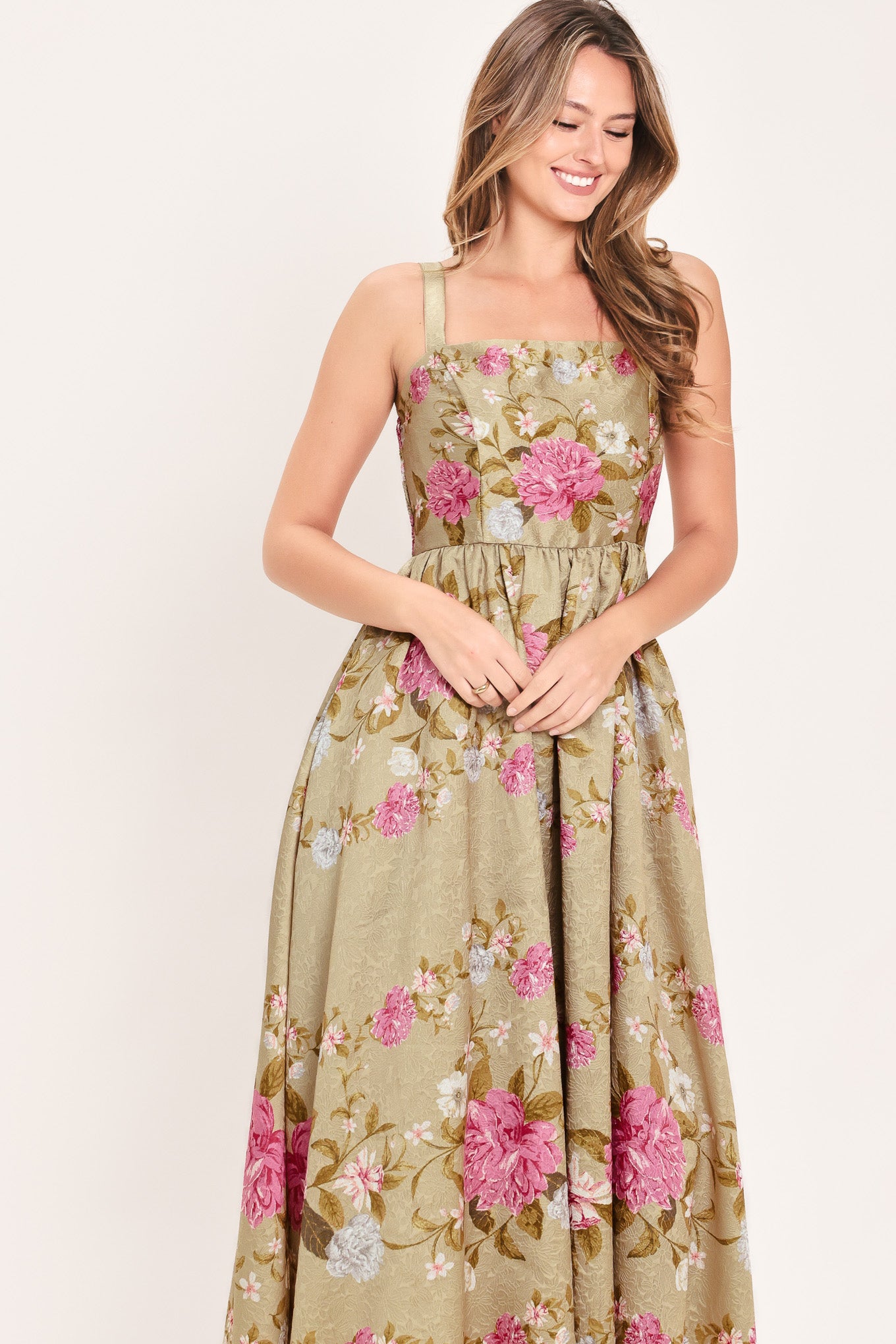 A woman wearing a sleeveless midi dress with a floral print on tan background. The dress features pink, white, and yellow flowers with green leaves | Flying Tomato