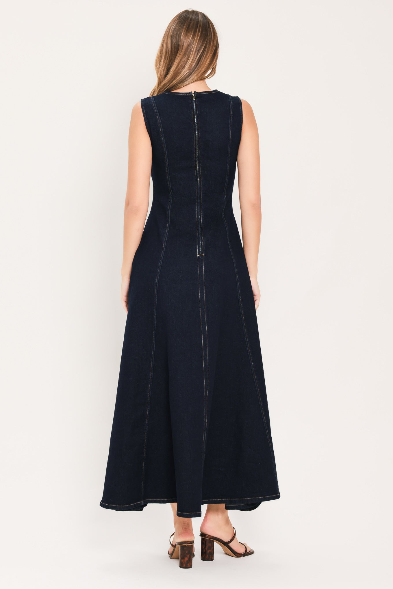 A woman wearing a sleeveless dark navy maxi dress with a crew neckline, a horizontal cutout across the chest, and small gold stud embellishments | Flying Tomato