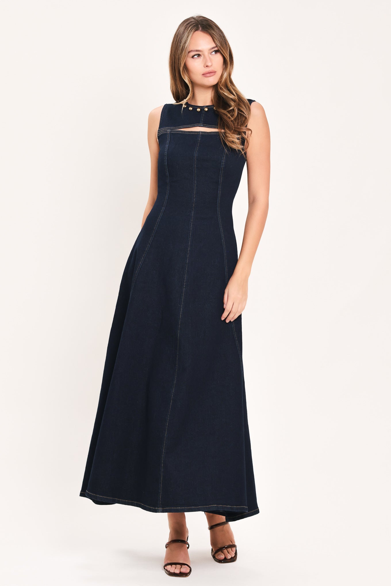 A woman wearing a sleeveless dark navy maxi dress with a crew neckline, a horizontal cutout across the chest, and small gold stud embellishments | Flying Tomato