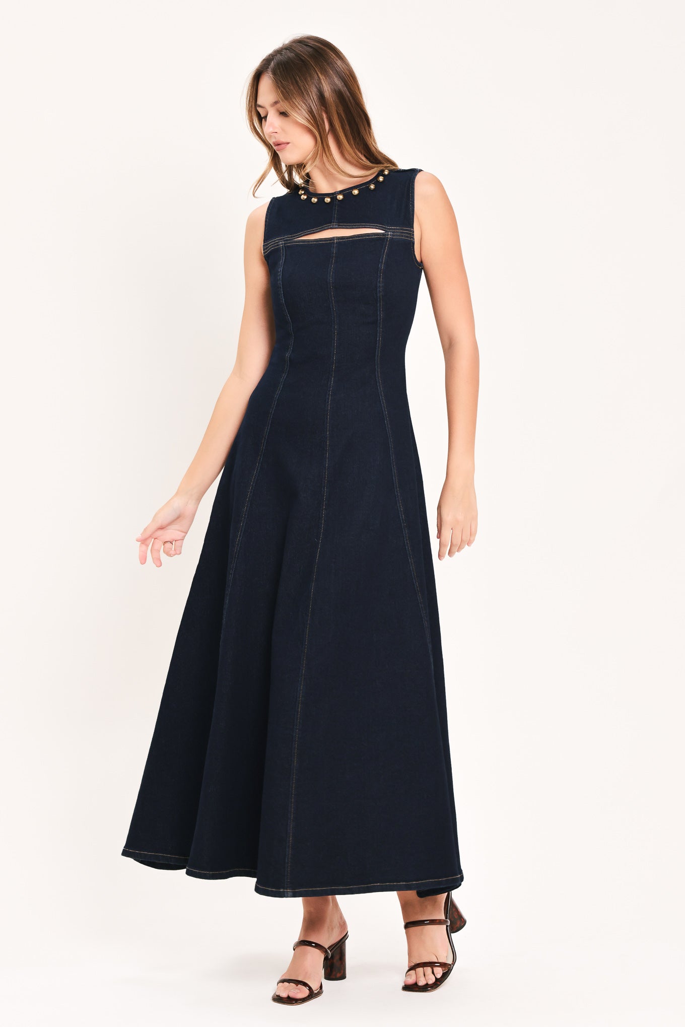 A woman wearing a sleeveless dark navy maxi dress with a crew neckline, a horizontal cutout across the chest, and small gold stud embellishments | Flying Tomato