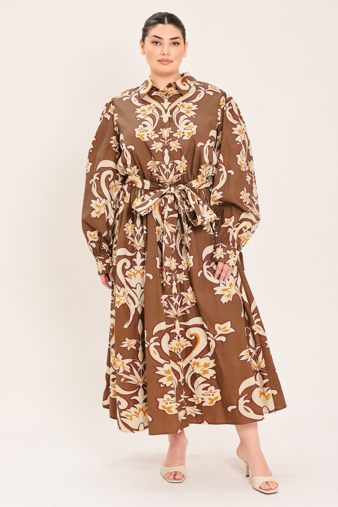 A woman modeling a brown long-sleeve midi dress with a cream and gold baroque-style floral pattern, featuring a tie waist belt and a shawl collar | Plus Curve Flying Tomato