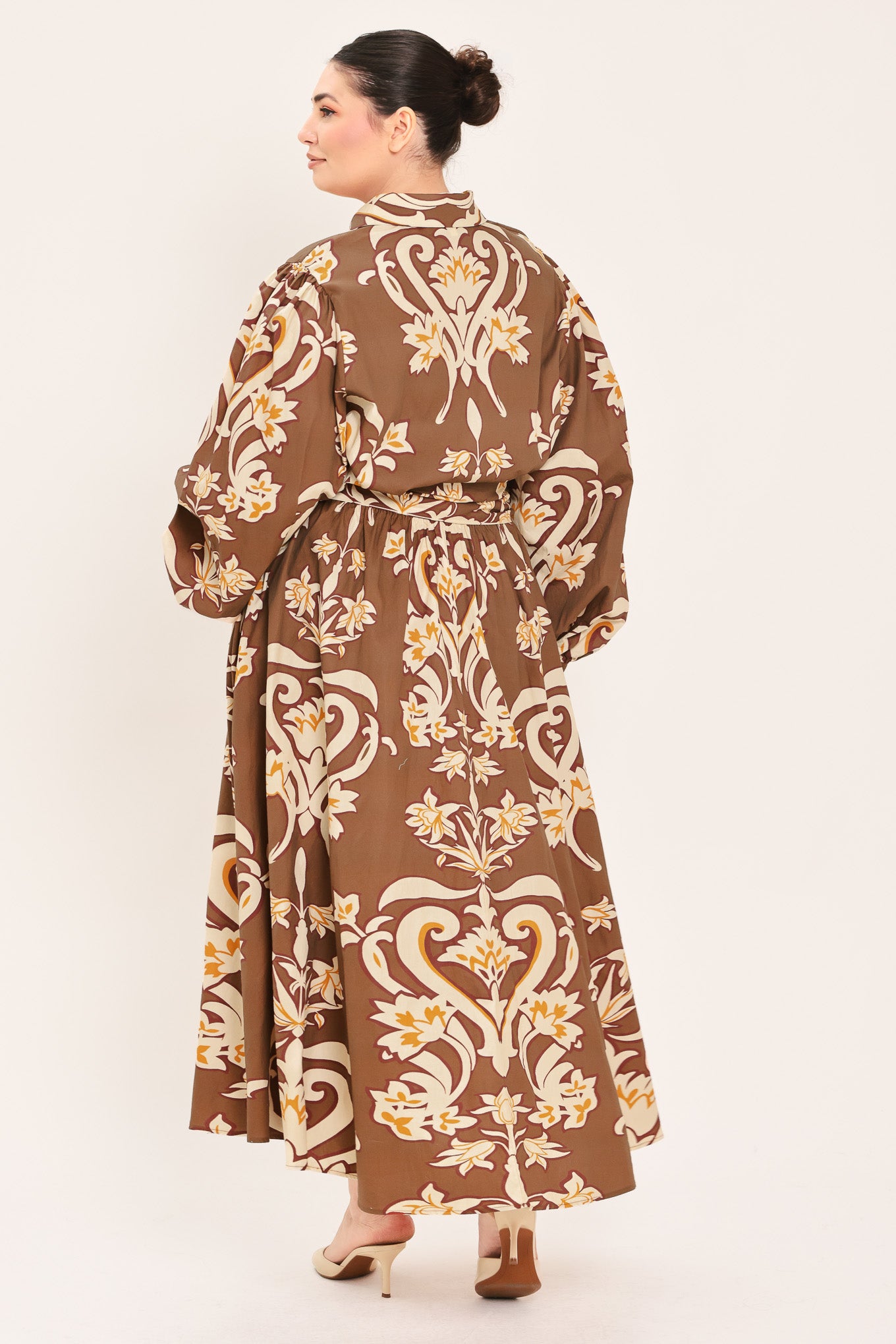 A woman modeling a brown long-sleeve midi dress with a cream and gold baroque-style floral pattern, featuring a tie waist belt and a shawl collar | Plus Curve Flying Tomato
