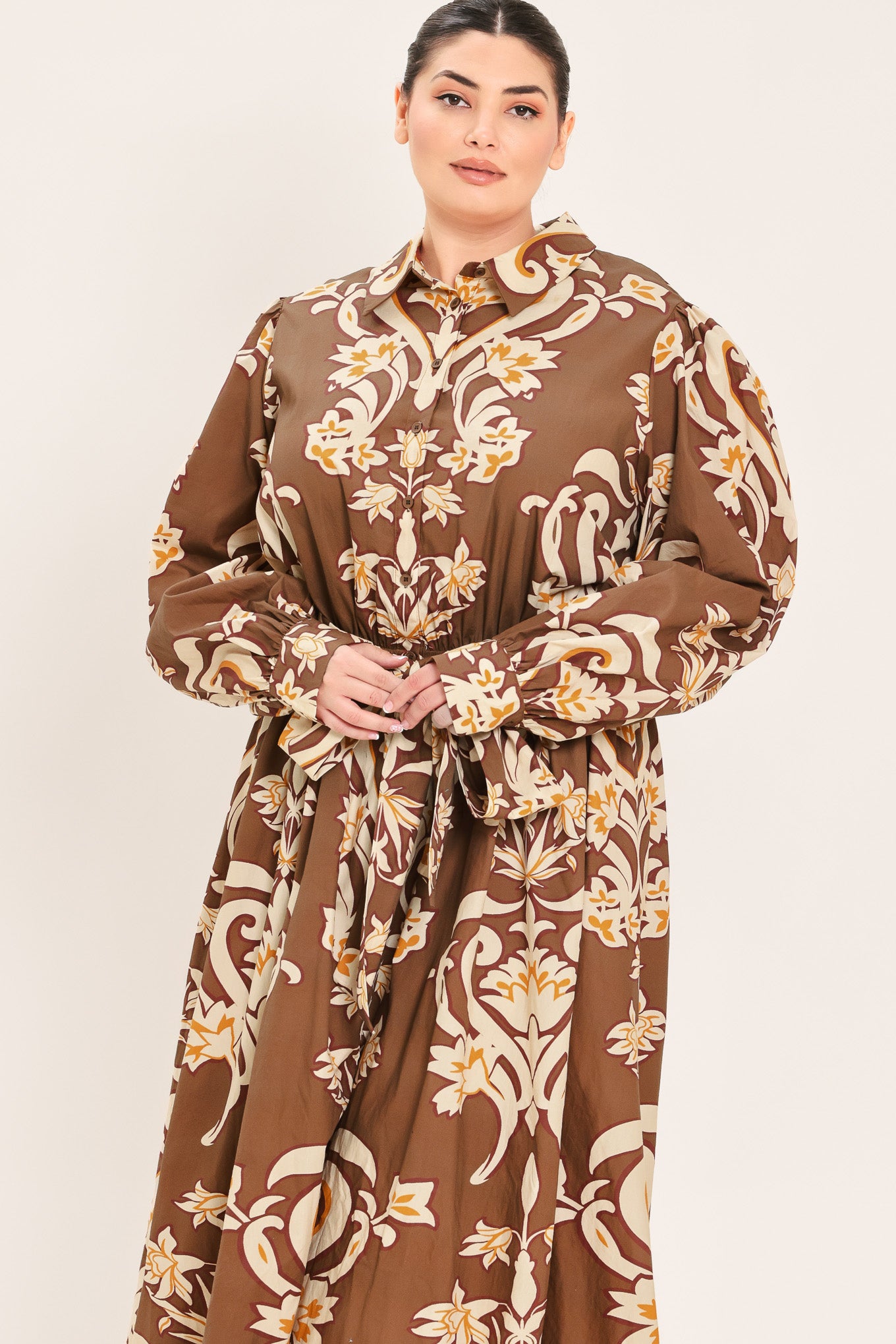 A woman modeling a brown long-sleeve midi dress with a cream and gold baroque-style floral pattern, featuring a tie waist belt and a shawl collar | Plus Curve Flying Tomato