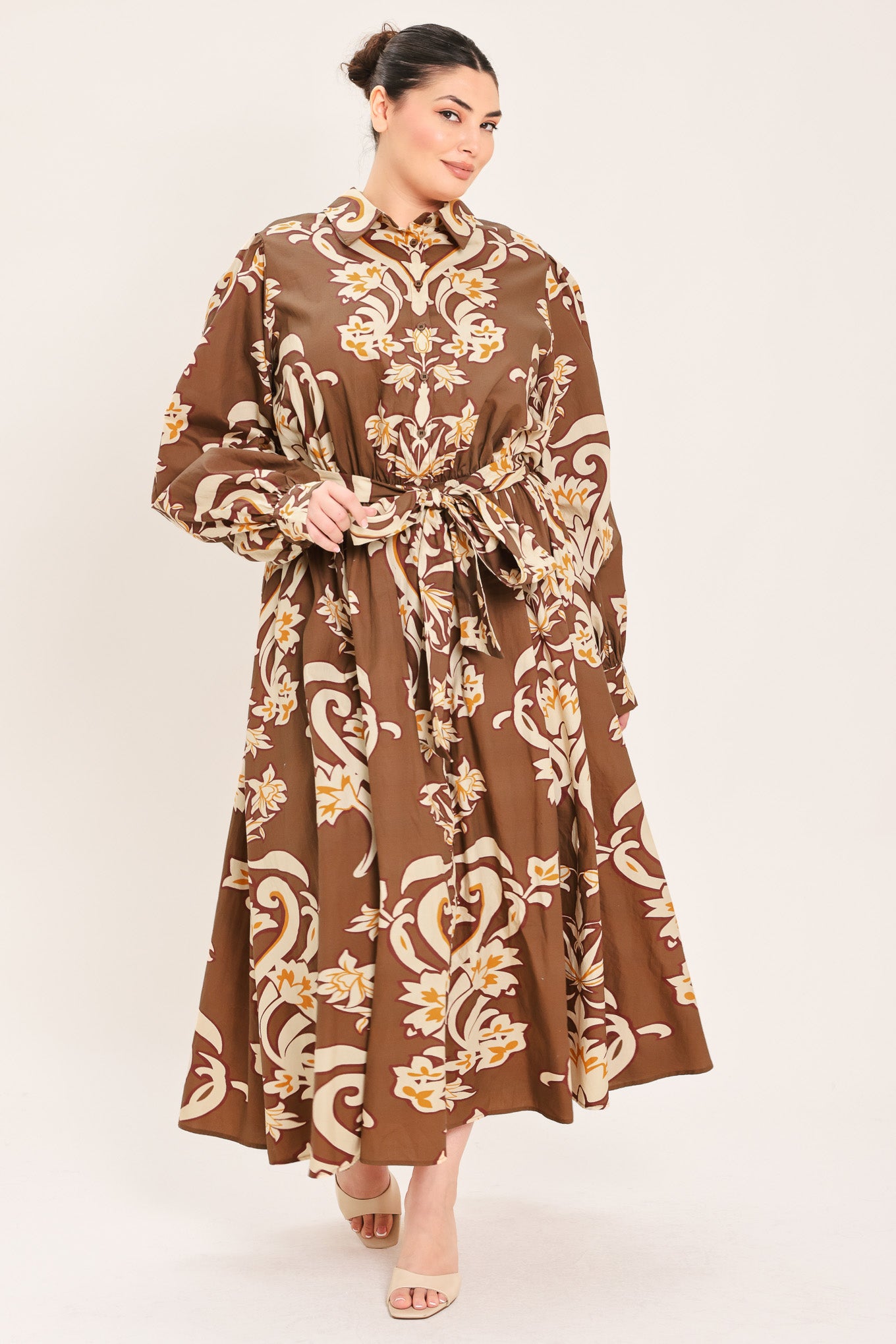 A woman modeling a brown long-sleeve midi dress with a cream and gold baroque-style floral pattern, featuring a tie waist belt and a shawl collar | Plus Curve Flying Tomato