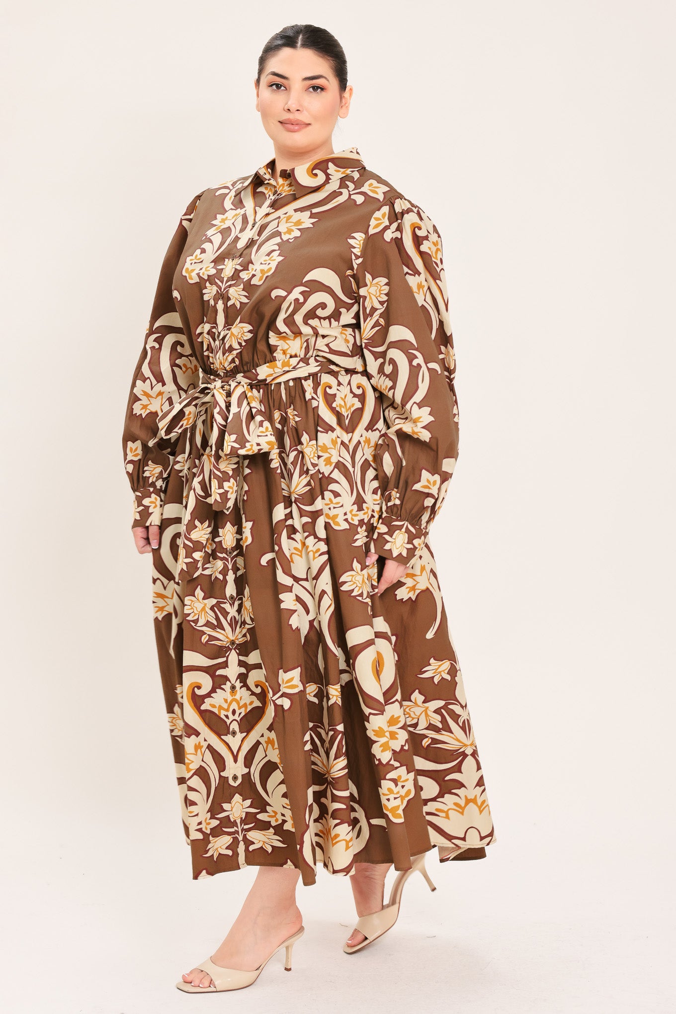 A woman modeling a brown long-sleeve midi dress with a cream and gold baroque-style floral pattern, featuring a tie waist belt and a shawl collar | Plus Curve Flying Tomato
