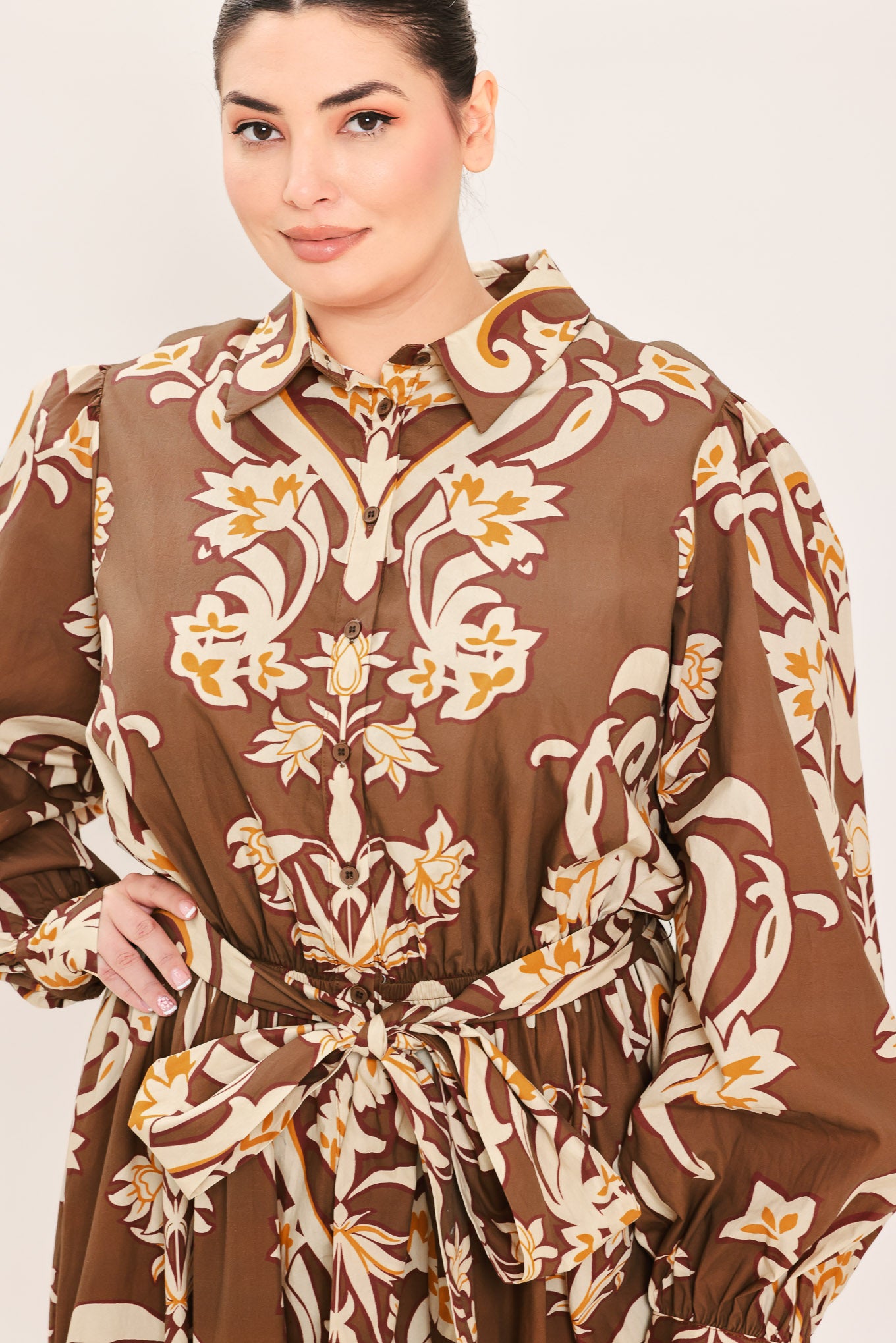A woman modeling a brown long-sleeve midi dress with a cream and gold baroque-style floral pattern, featuring a tie waist belt and a shawl collar | Plus Curve Flying Tomato